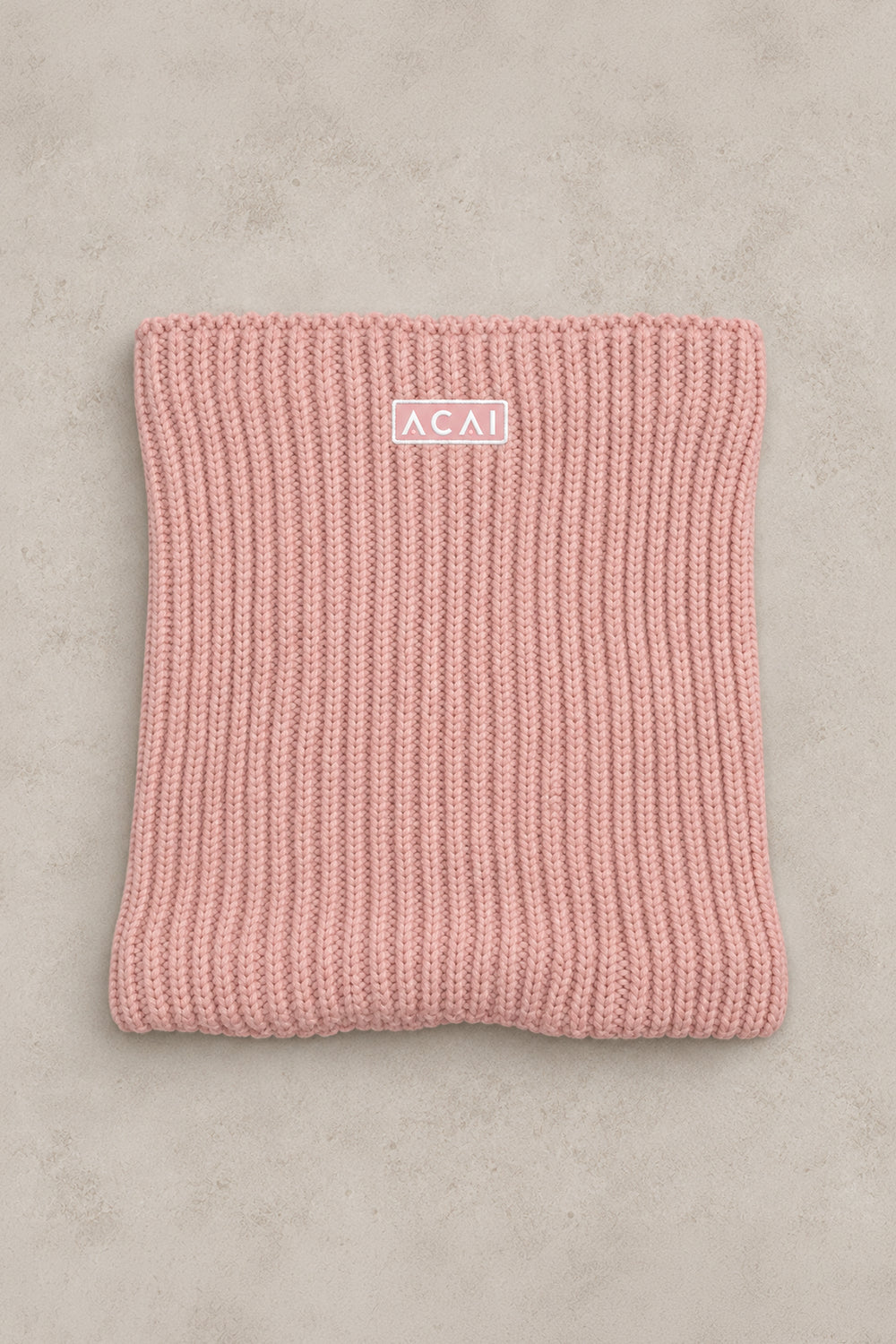 A folded dusty pink ribbed knit beanie with a small white rectangular logo patch reading 'ACAI' on a textured light gray background.