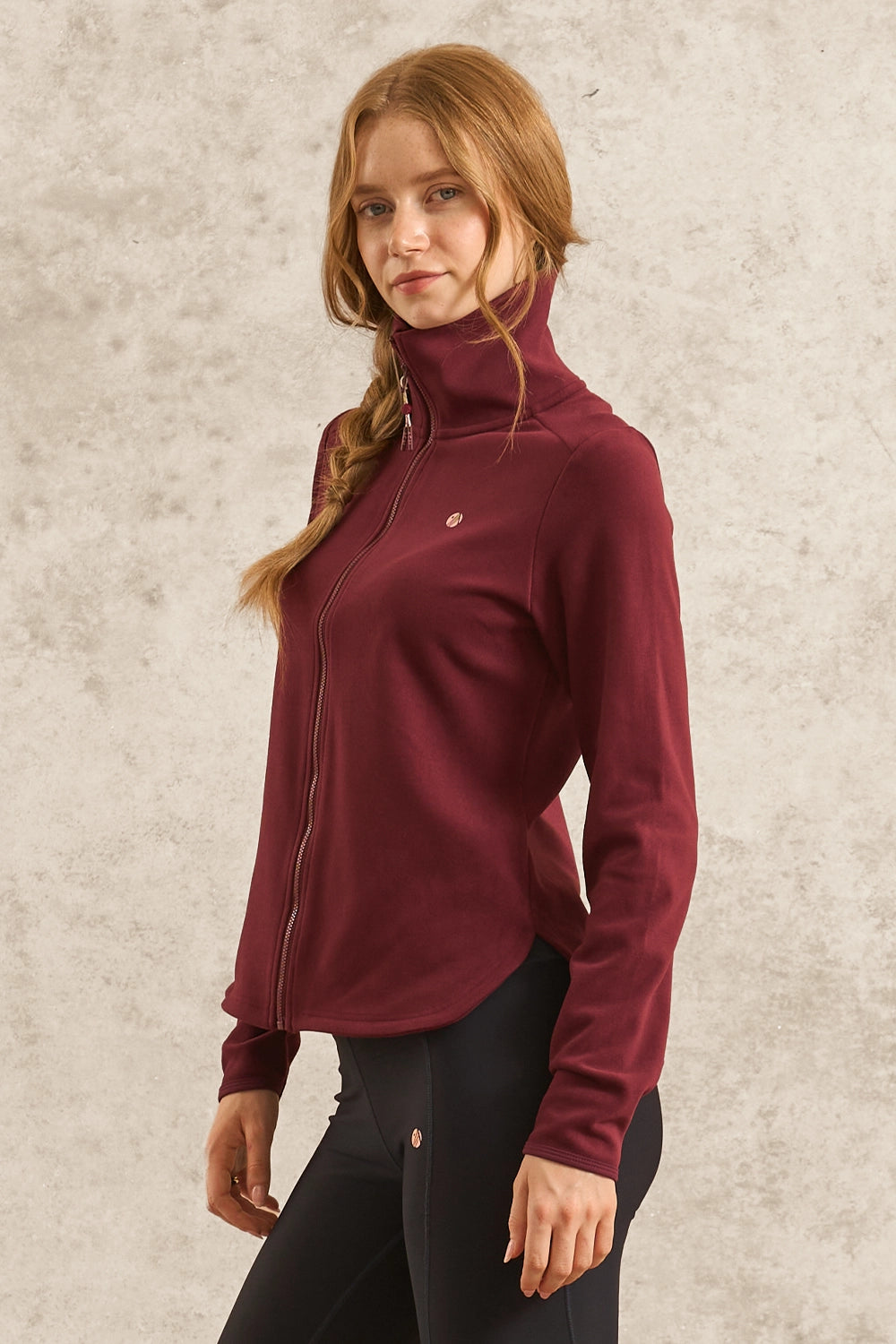 A woman wearing a burgundy long-sleeve zip-up athletic jacket with a mock neck collar and black leggings, standing against a textured light gray wall.