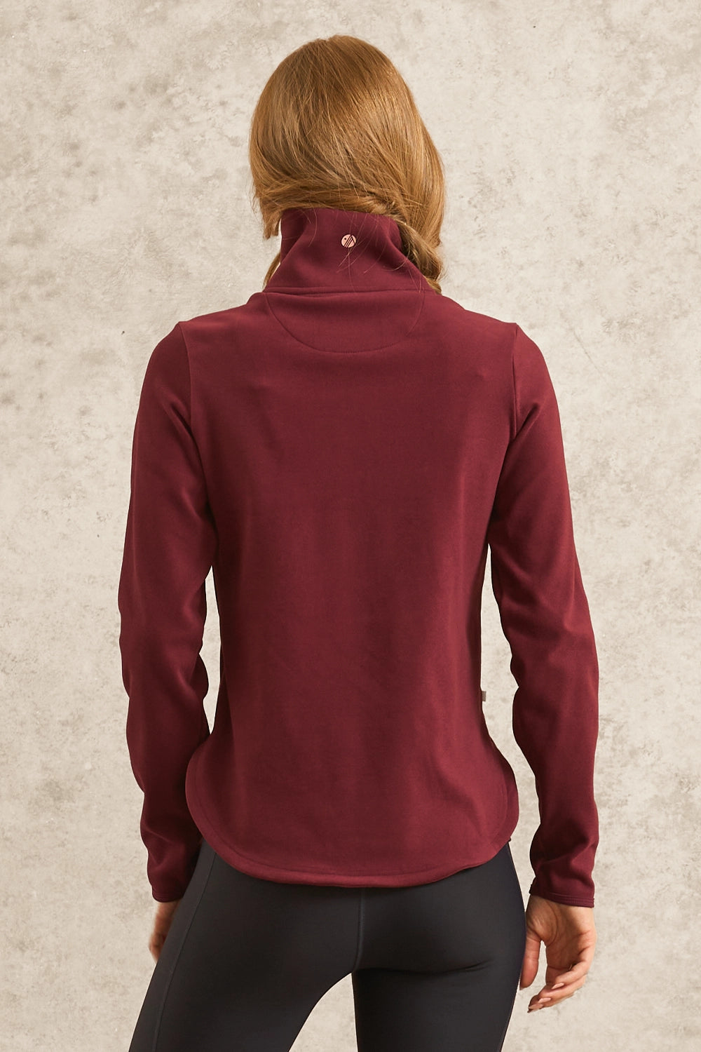 A woman viewed from the back wearing a long-sleeve high-neck or mock-neck athletic top in deep burgundy, paired with black leggings, against a textured light gray wall.