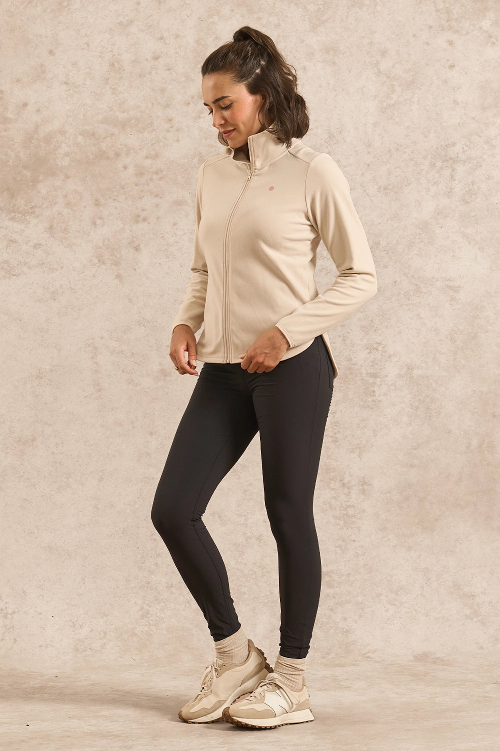 A woman modeling a beige long-sleeve athletic quarter-zip jacket and black leggings, standing against a textured beige studio backdrop.