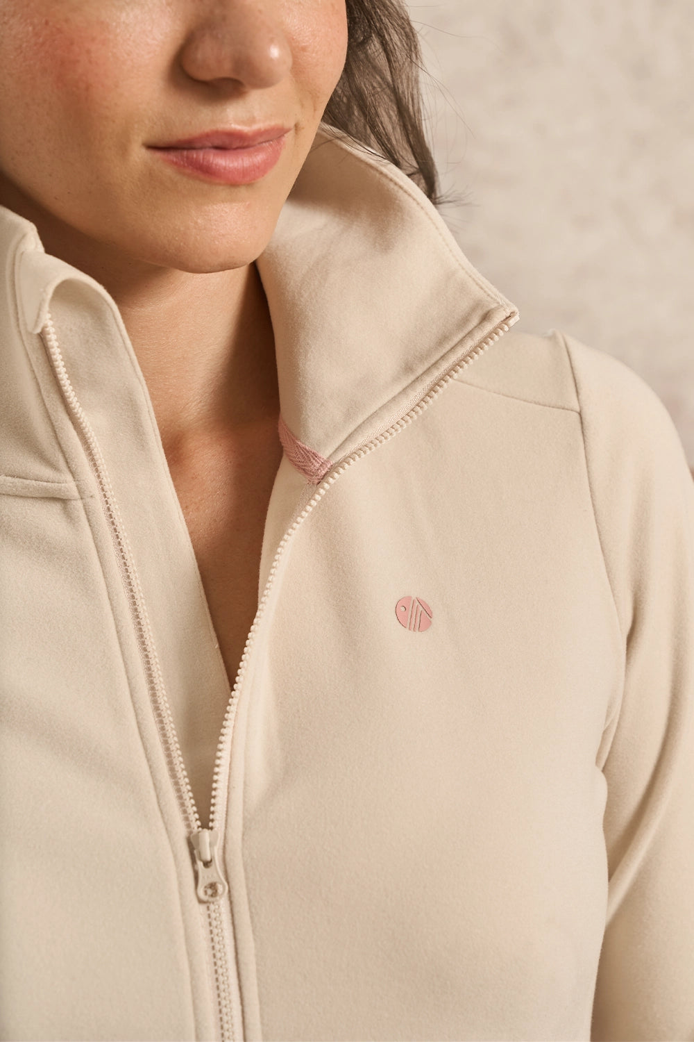 Close-up of a person wearing a light beige zip-up fleece jacket with a small pink circular logo on the chest.