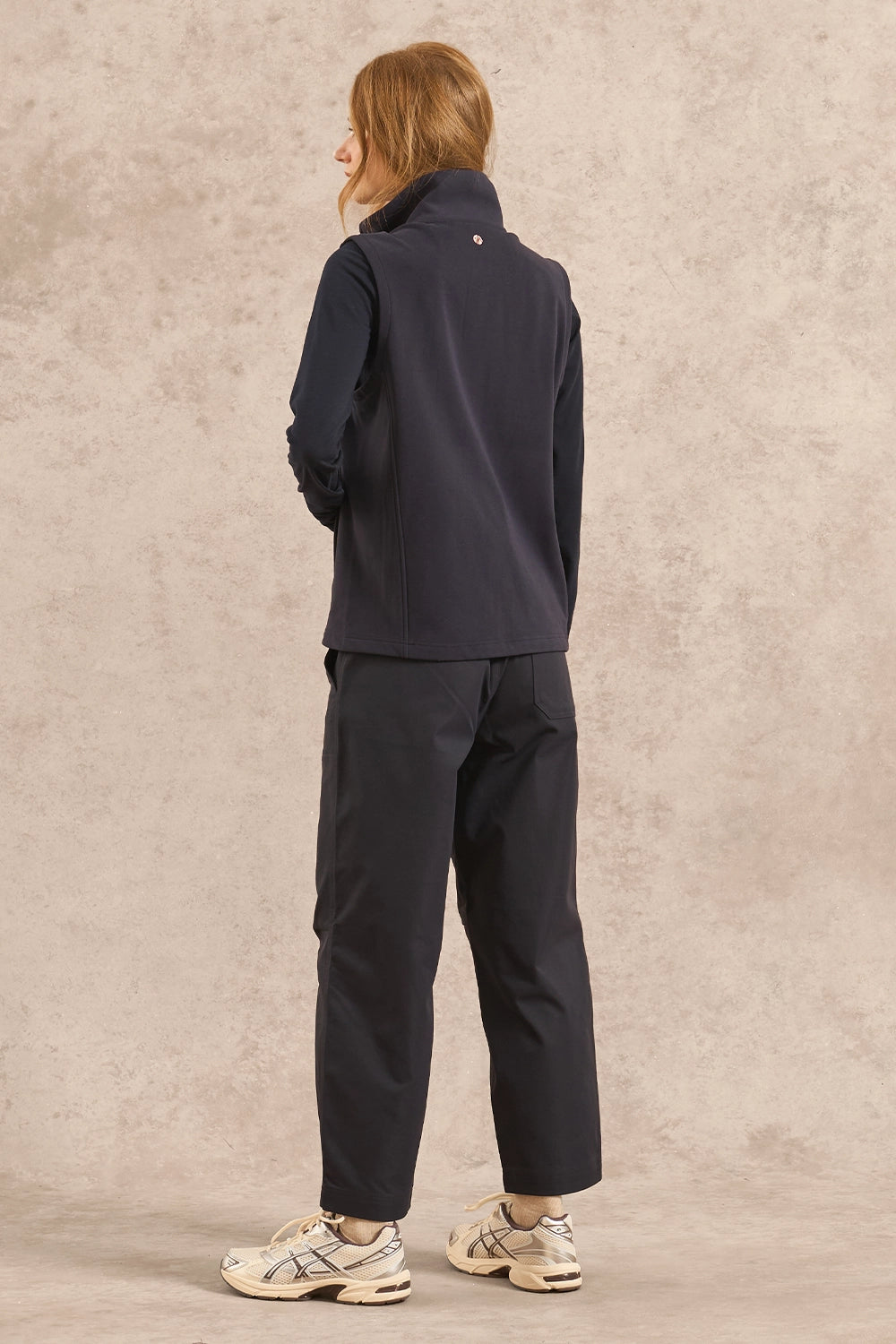 A model is shown from the back wearing a dark navy fleece vest and matching dark navy pants, paired with beige sneakers, against a textured beige studio backdrop.