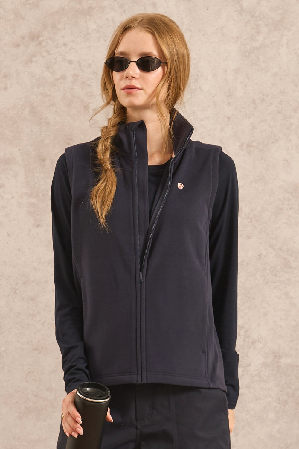 A woman wearing a dark navy zip-up fleece vest over a black top, styled with black sunglasses and holding a black travel tumbler against a textured beige background.