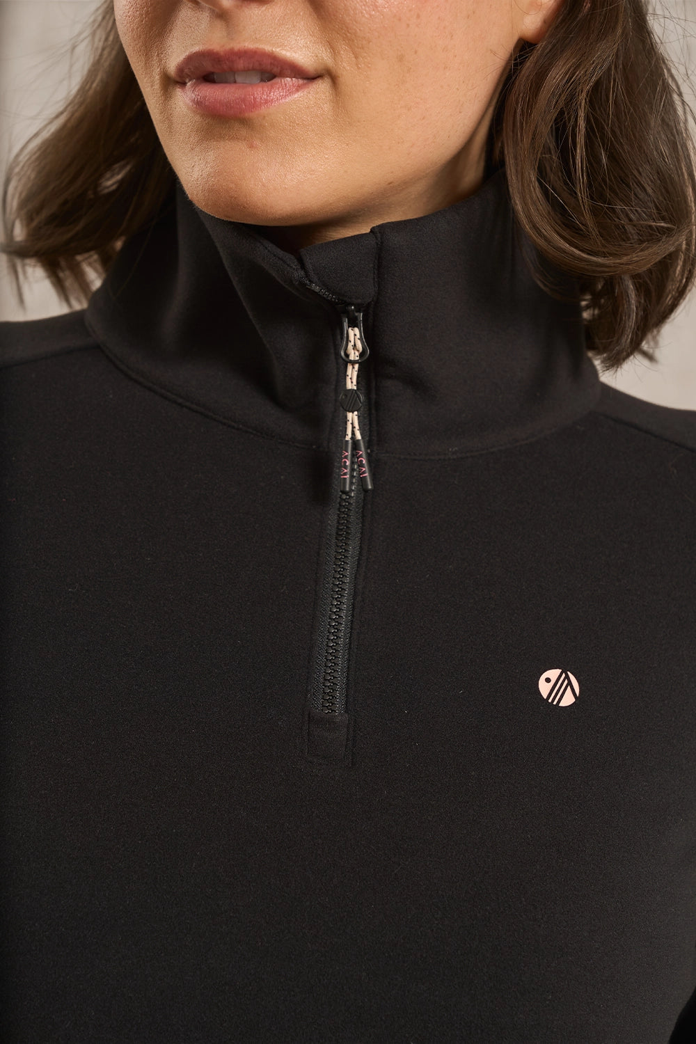 Close-up of a woman wearing a black quarter-zip pullover with a small pink circular logo on the left chest.