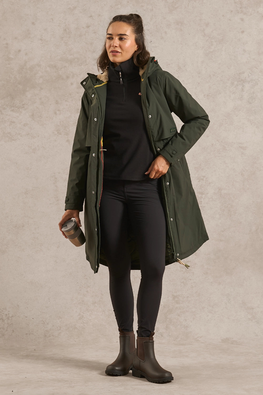 A woman modeling a mid-length olive green hooded raincoat over a black outfit, standing against a textured light gray studio backdrop and holding a small cup.
