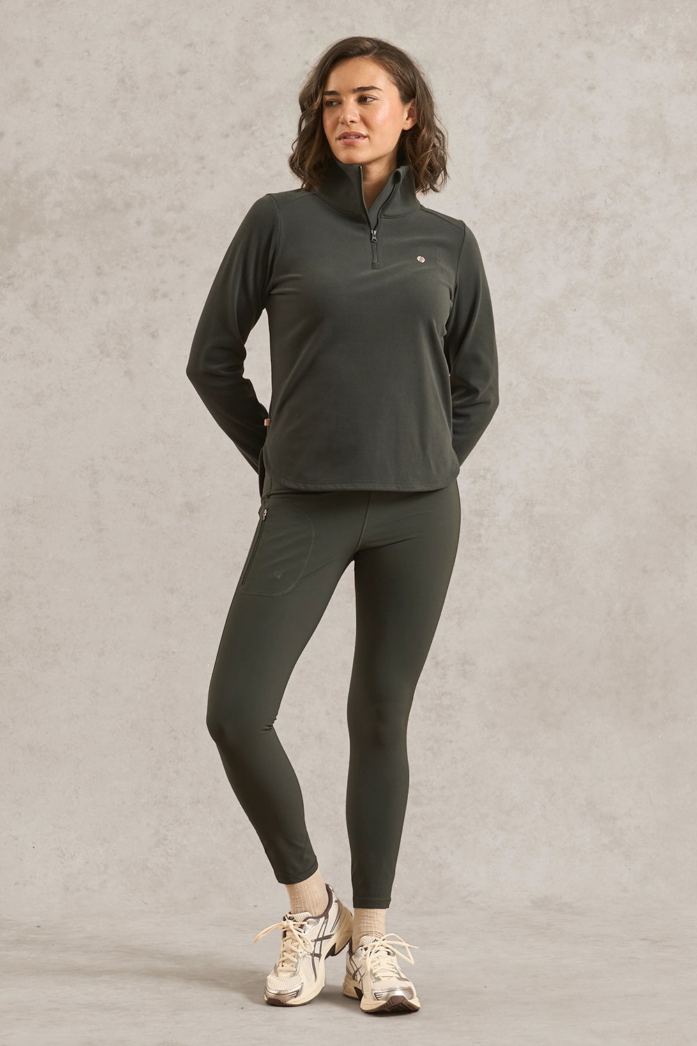 A woman modeling a dark olive green athletic set consisting of a quarter-zip long-sleeve top and matching leggings, paired with white sneakers, against a textured light gray studio backdrop.