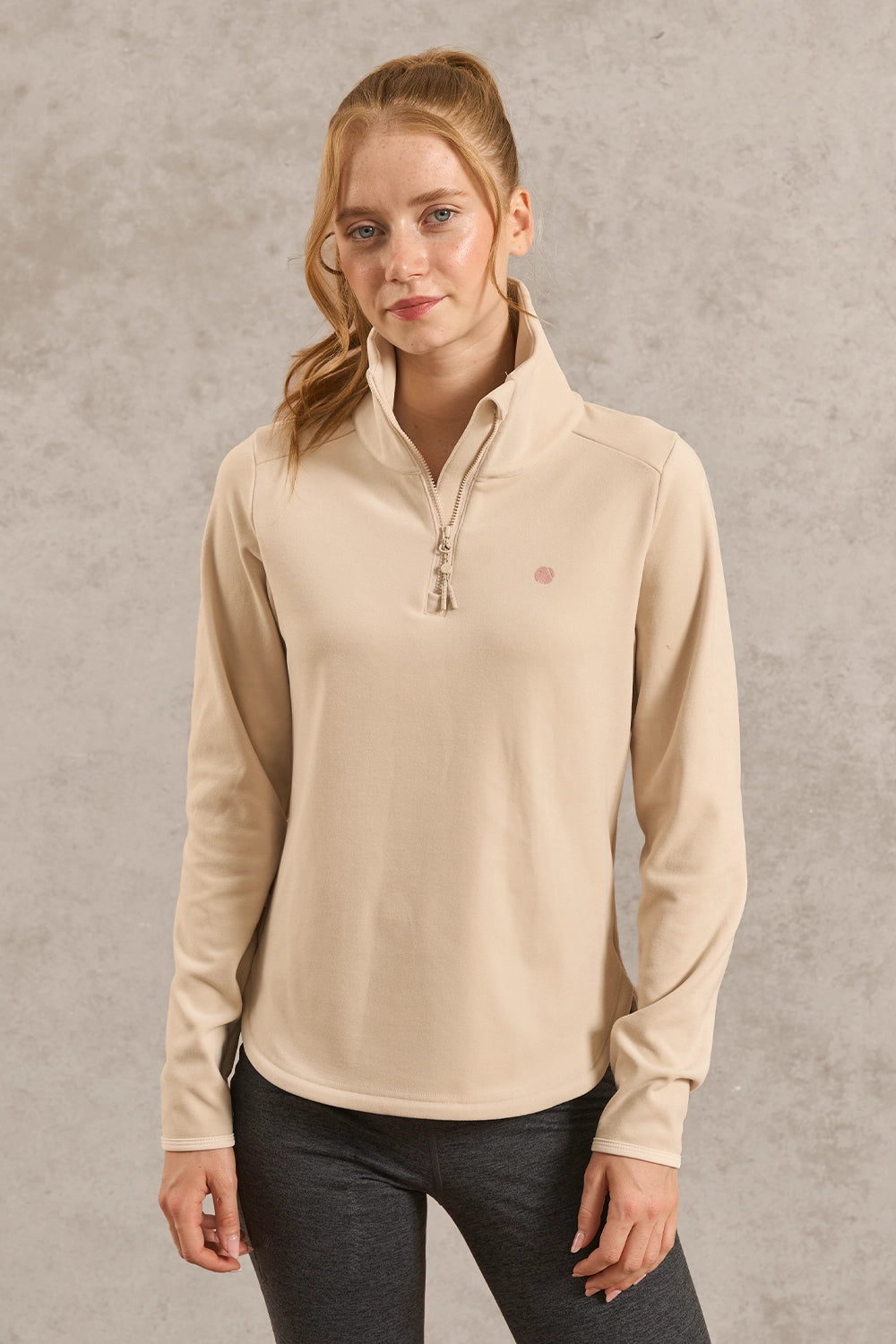 A woman modeling a beige long-sleeve quarter-zip pullover with a small pink embroidered logo on the left chest, paired with dark gray leggings, against a textured gray studio background.