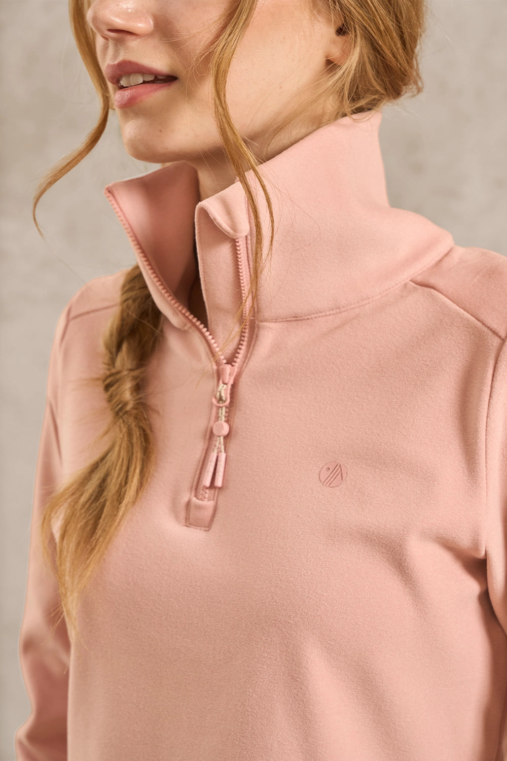 A close-up of a woman wearing a dusty pink quarter-zip pullover sweatshirt with a high collar and a small tonal logo on the chest.