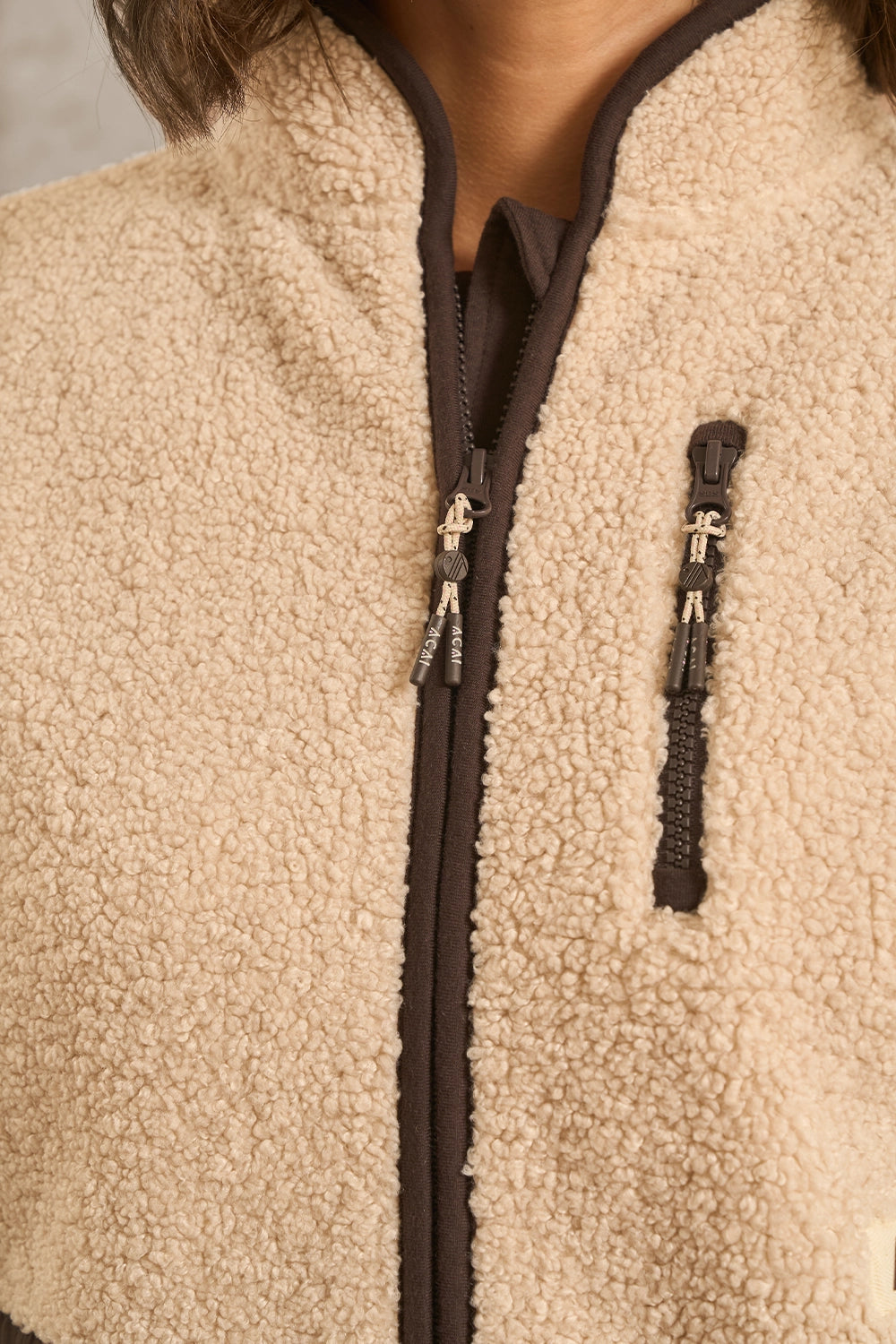 Close-up of a beige sherpa fleece zip-up jacket with dark brown trim, zipper, and zipper pulls, worn by a model.