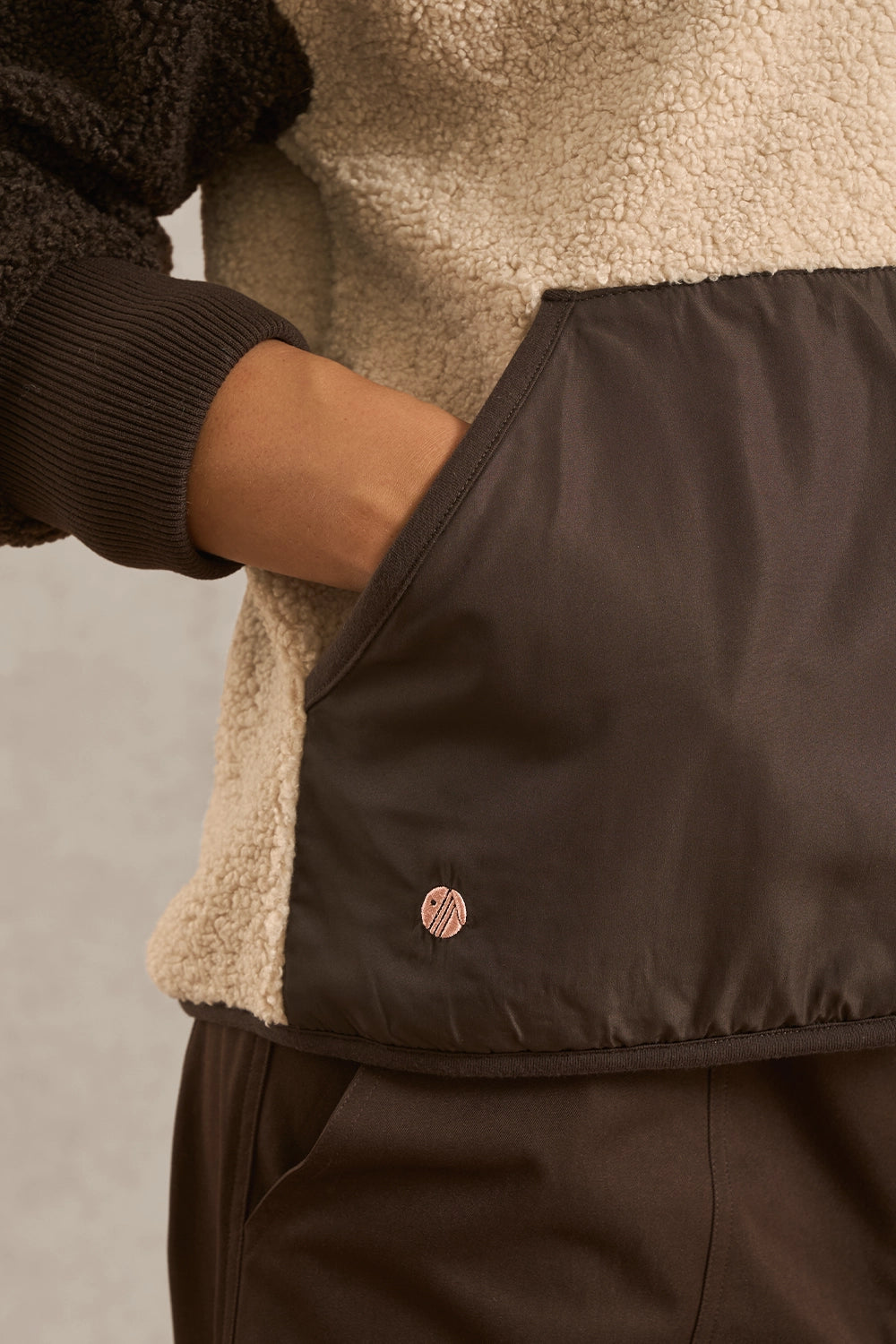 Close-up of a person wearing a dark brown jacket with a beige sherpa lining and a matching dark brown skirt or pants, showing a small circular metallic logo on the jacket pocket.
