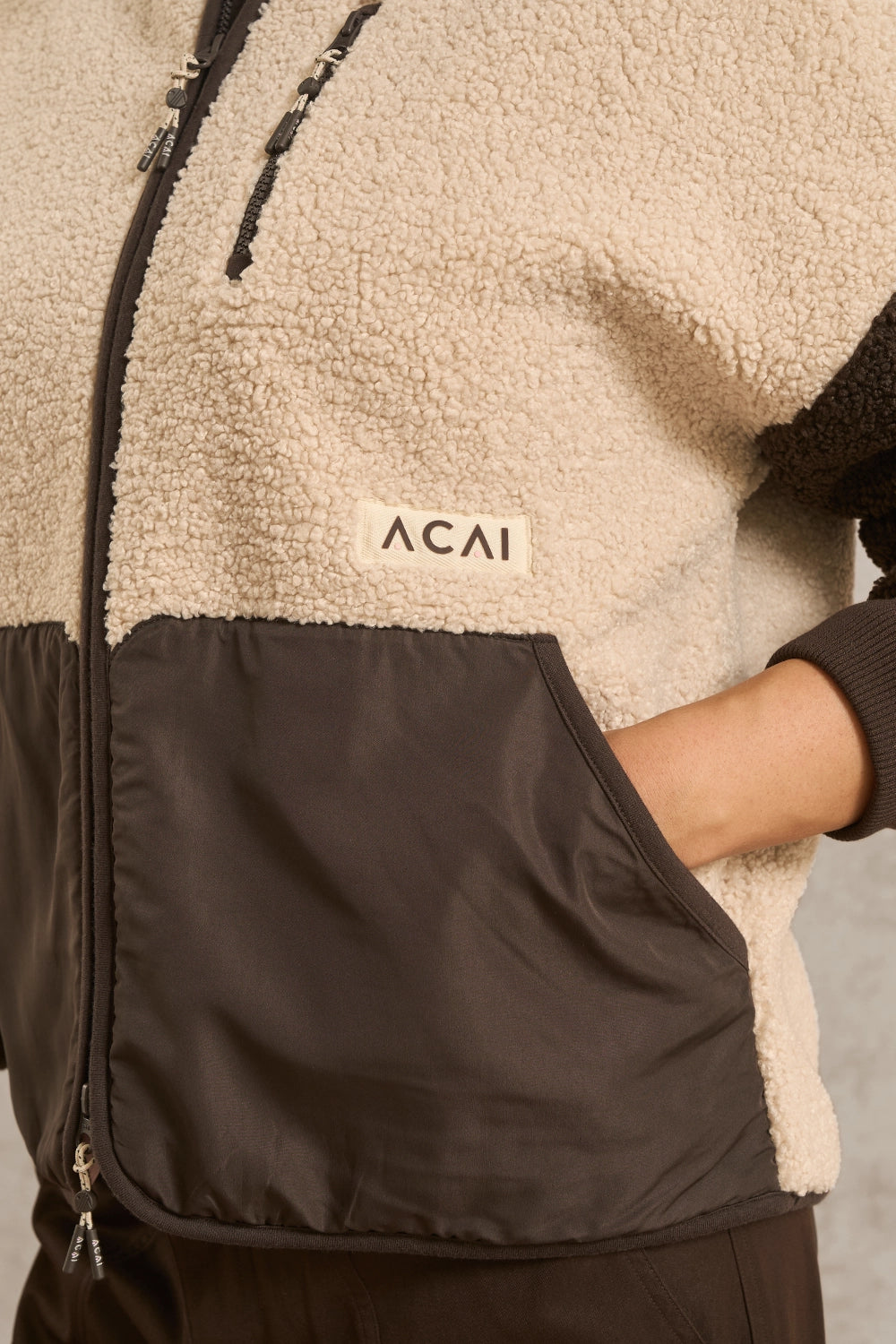 Close-up of a person wearing a fleece zip jacket with a cream sherpa upper section, dark brown lower panel, and a small white ACAI logo patch on the chest.