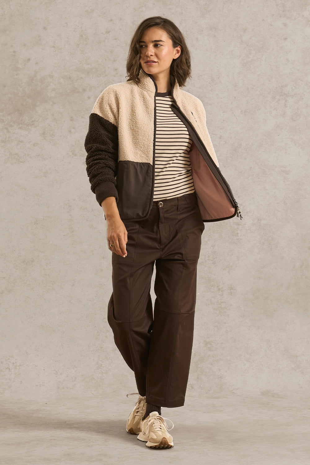 A woman models a color-block fleece jacket with a cream sherpa upper section, dark brown sleeves and side panels, and a brown mesh-lined front. She is styled with a black and white striped t-shirt, dark brown wide-leg cargo pants, and beige sneakers, set against a textured light gray studio backdrop.