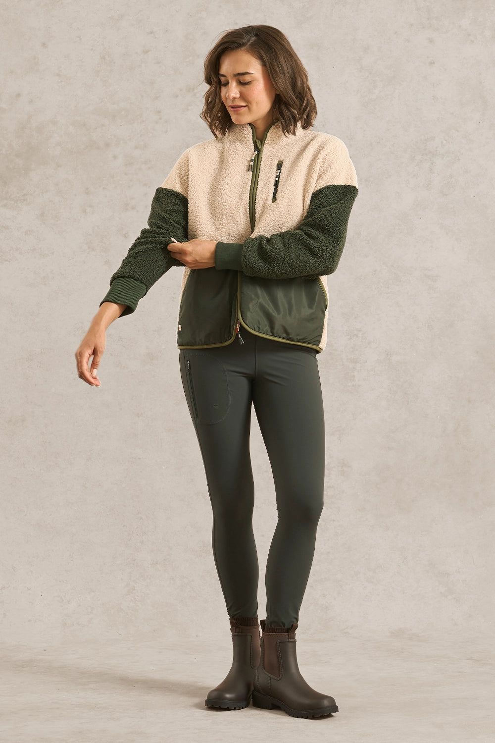 A woman modeling a color-block fleece jacket with a cream sherpa upper section, dark green sleeves, and dark green lower panels, paired with dark green leggings and dark brown boots, against a textured light gray studio backdrop.