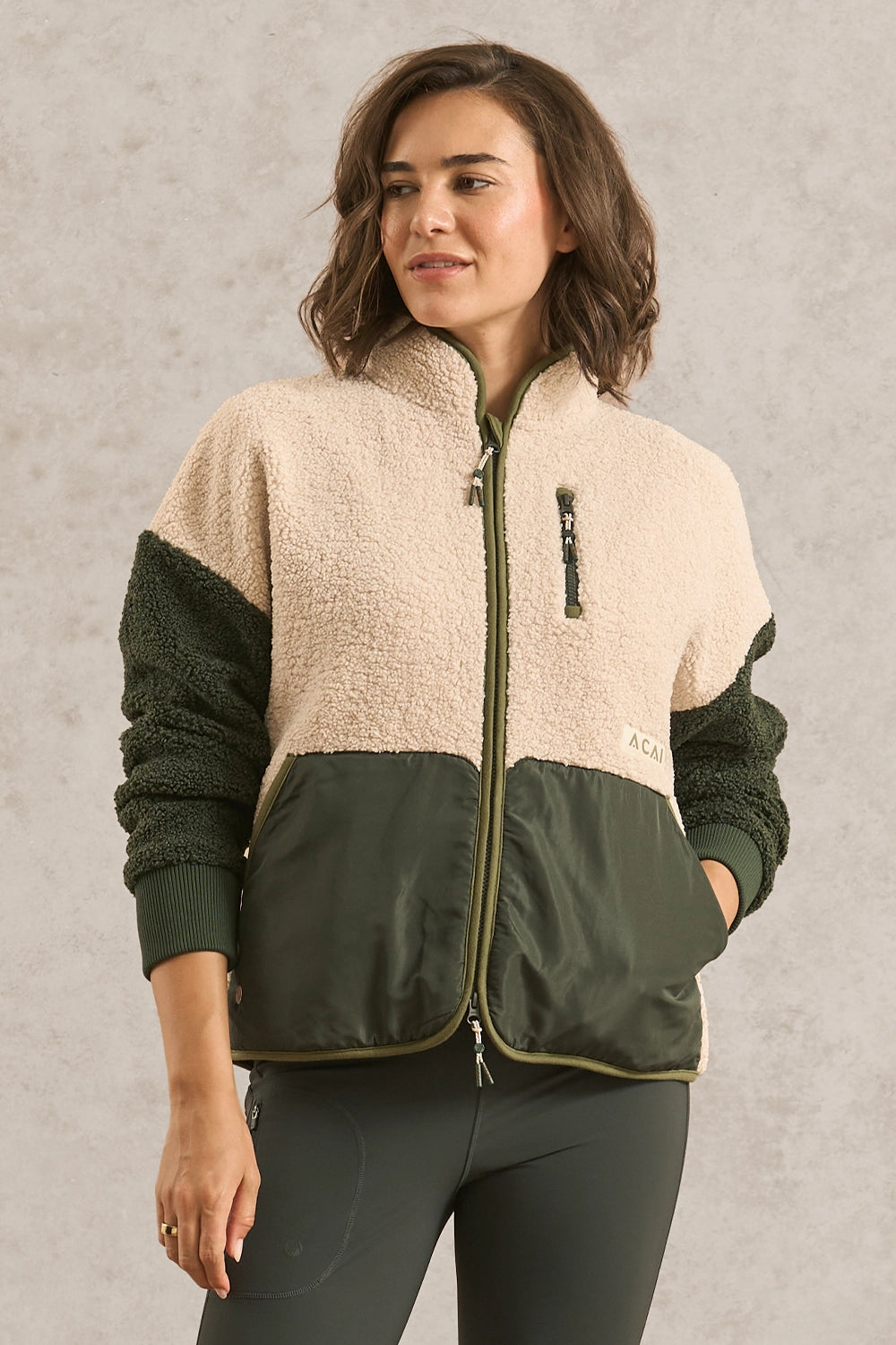 A woman wearing a color-block fleece jacket with a cream sherpa upper section, dark green sleeves, and a dark green front panel, paired with dark green leggings, set against a textured gray studio background.
