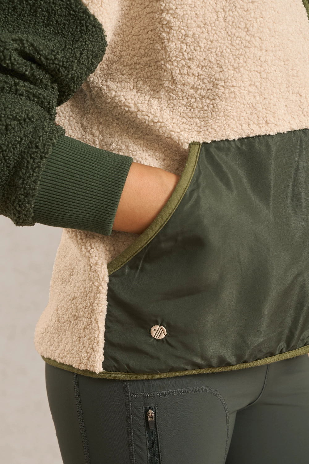 Close-up of a person wearing a fleece jacket with a color-blocked design, featuring a cream sherpa lining, dark green ribbed cuffs, and an olive green nylon front pocket with a small circular logo.