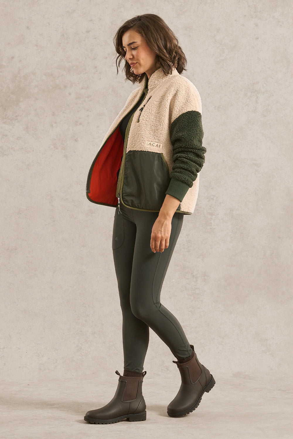A woman modeling a color-block fleece jacket with a cream sherpa upper, dark green sleeves and front panel, and a red interior lining, paired with dark green leggings and brown boots against a textured light gray studio backdrop.