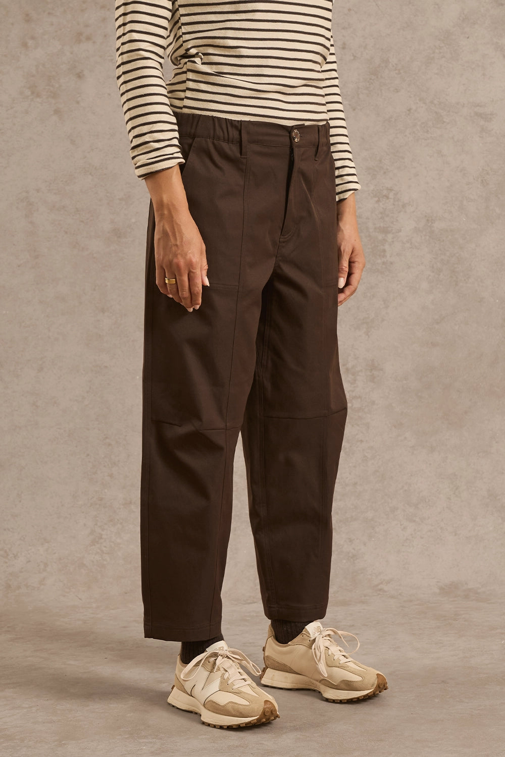 A person wearing dark brown wide-leg cargo-style pants, a cream and black striped long-sleeve top, and beige sneakers, standing against a textured gray studio backdrop.