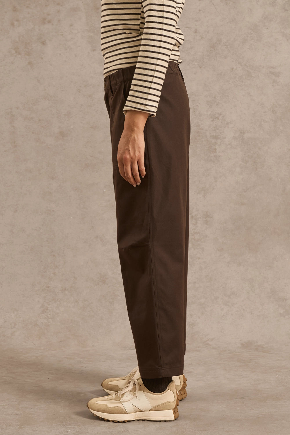 A person shown from the waist down wearing dark brown straight-leg trousers, a beige and black striped long-sleeve top, and beige sneakers, standing against a textured beige studio backdrop.
