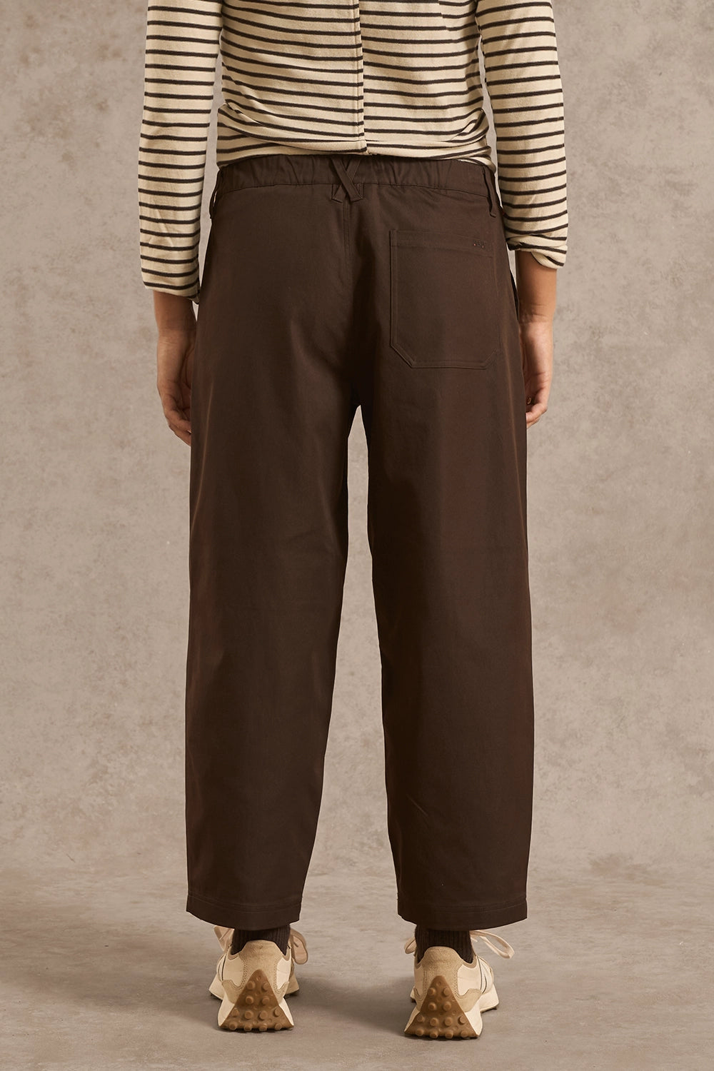 A person shown from the back wearing dark brown wide-leg trousers, a beige and black striped long-sleeve top, and beige sneakers against a textured beige studio backdrop.