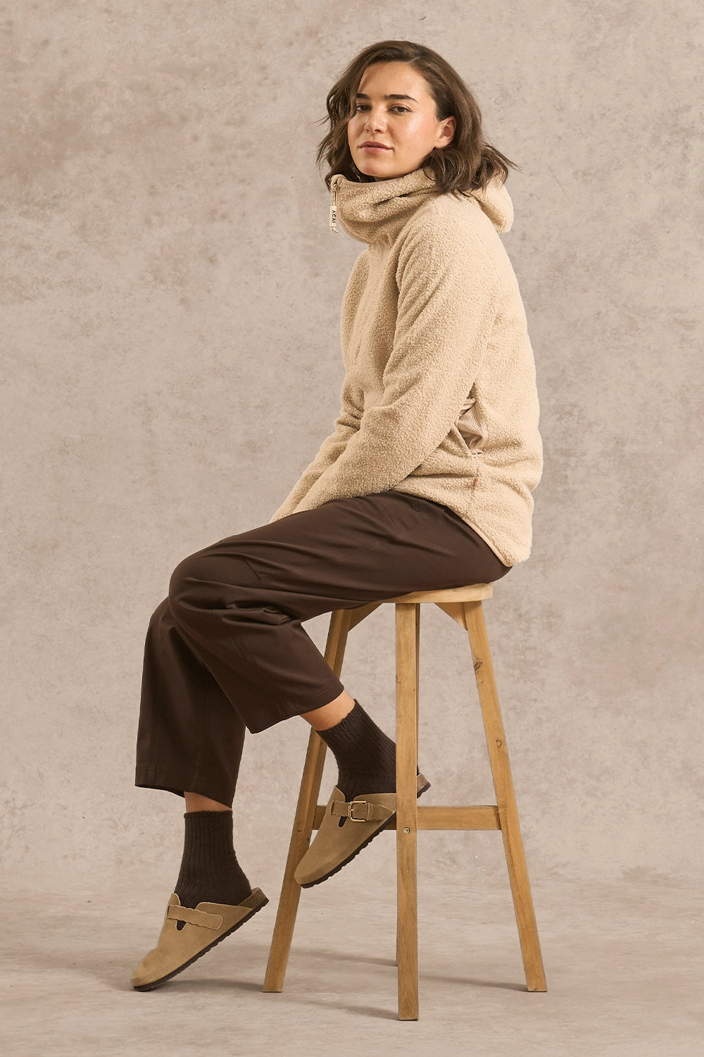 A woman sits on a wooden stool wearing a beige sherpa hoodie with a high neck, dark brown cropped pants, dark socks, and tan suede moccasin-style shoes against a textured beige studio backdrop.