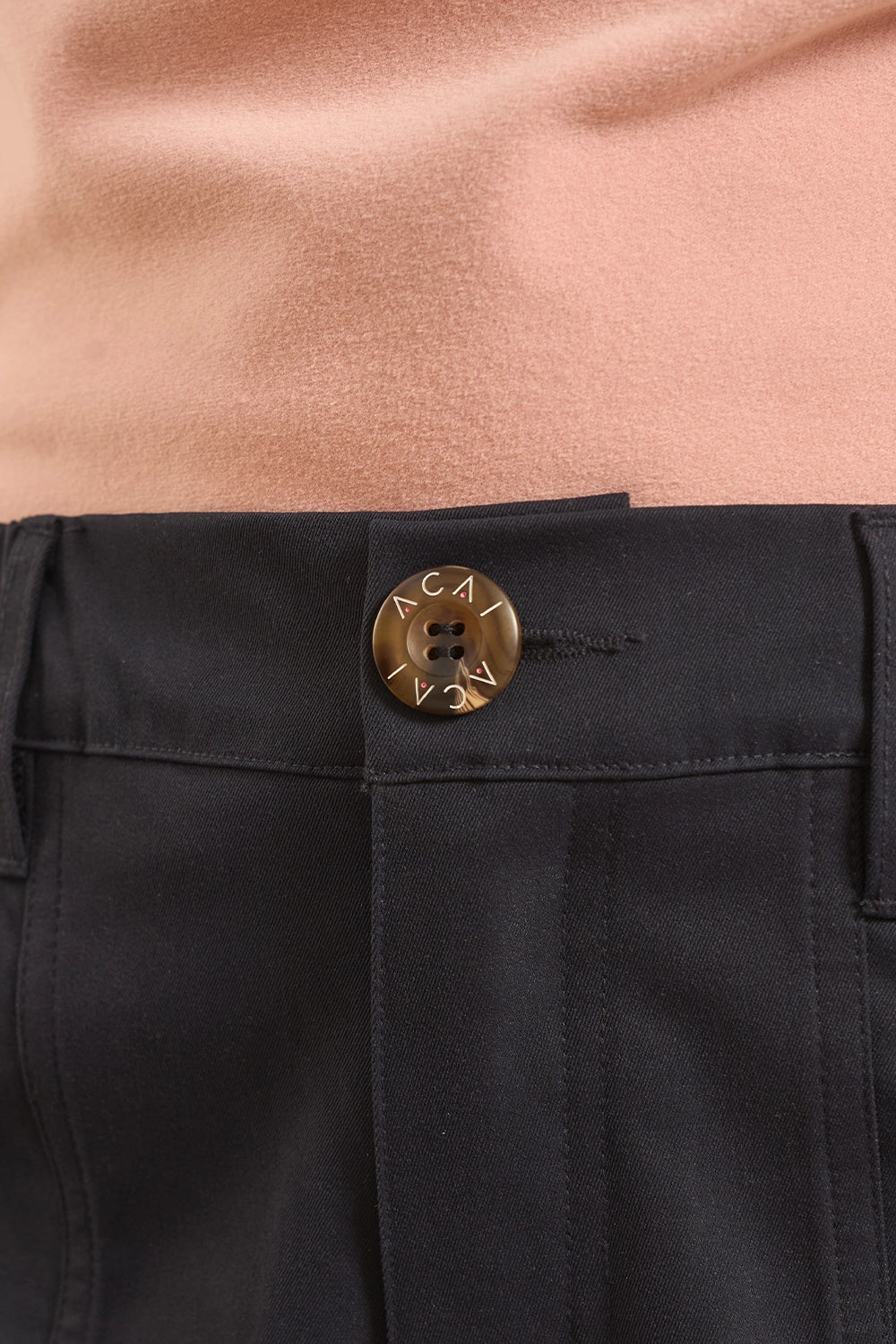Close-up of a black trouser waistband with a round gold-tone button engraved with the brand name 'ACAI', worn with a peach-colored top.