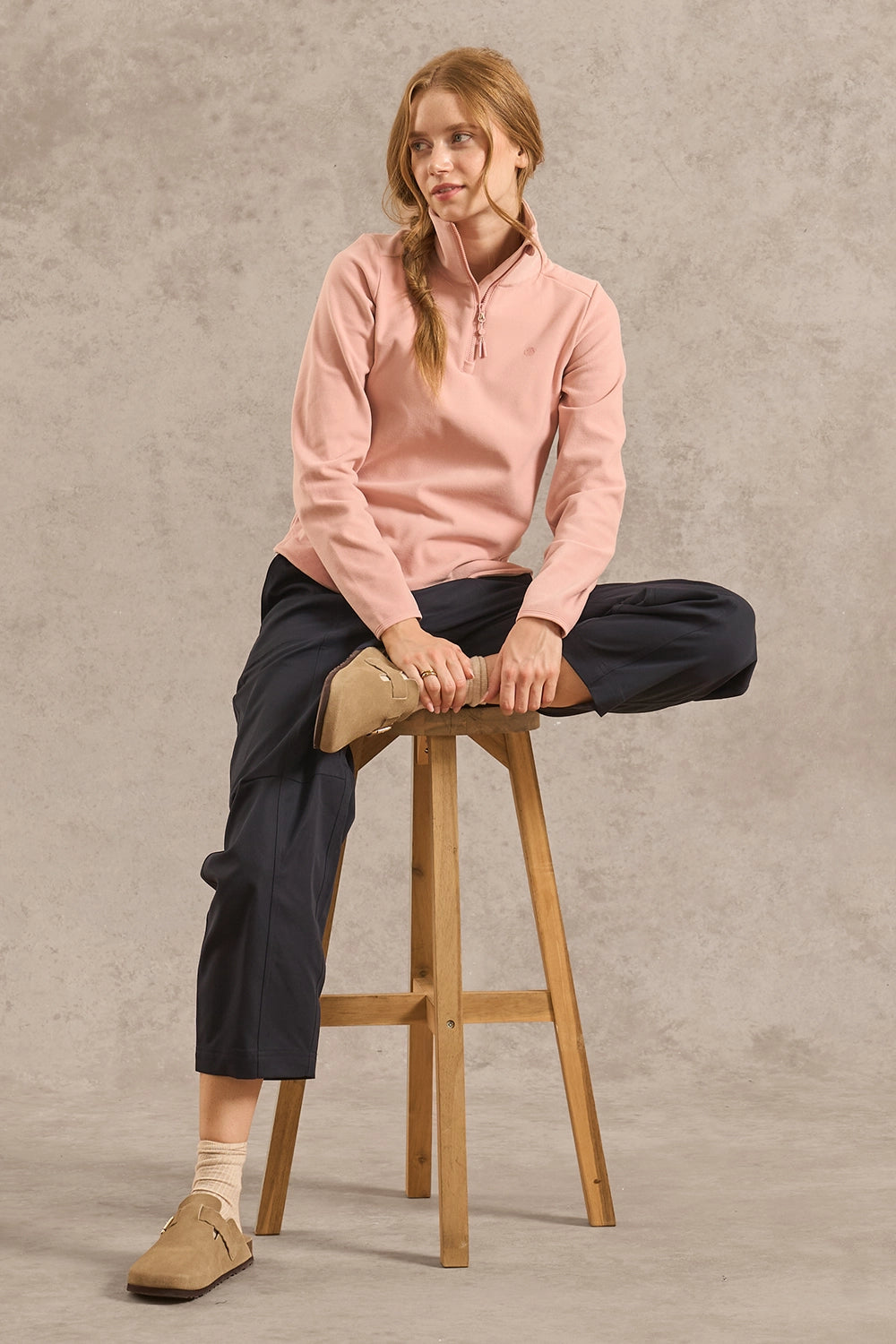 A woman sits on a wooden stool wearing a dusty pink quarter-zip long-sleeve top, dark navy cropped trousers, and tan suede loafers against a textured gray studio backdrop.