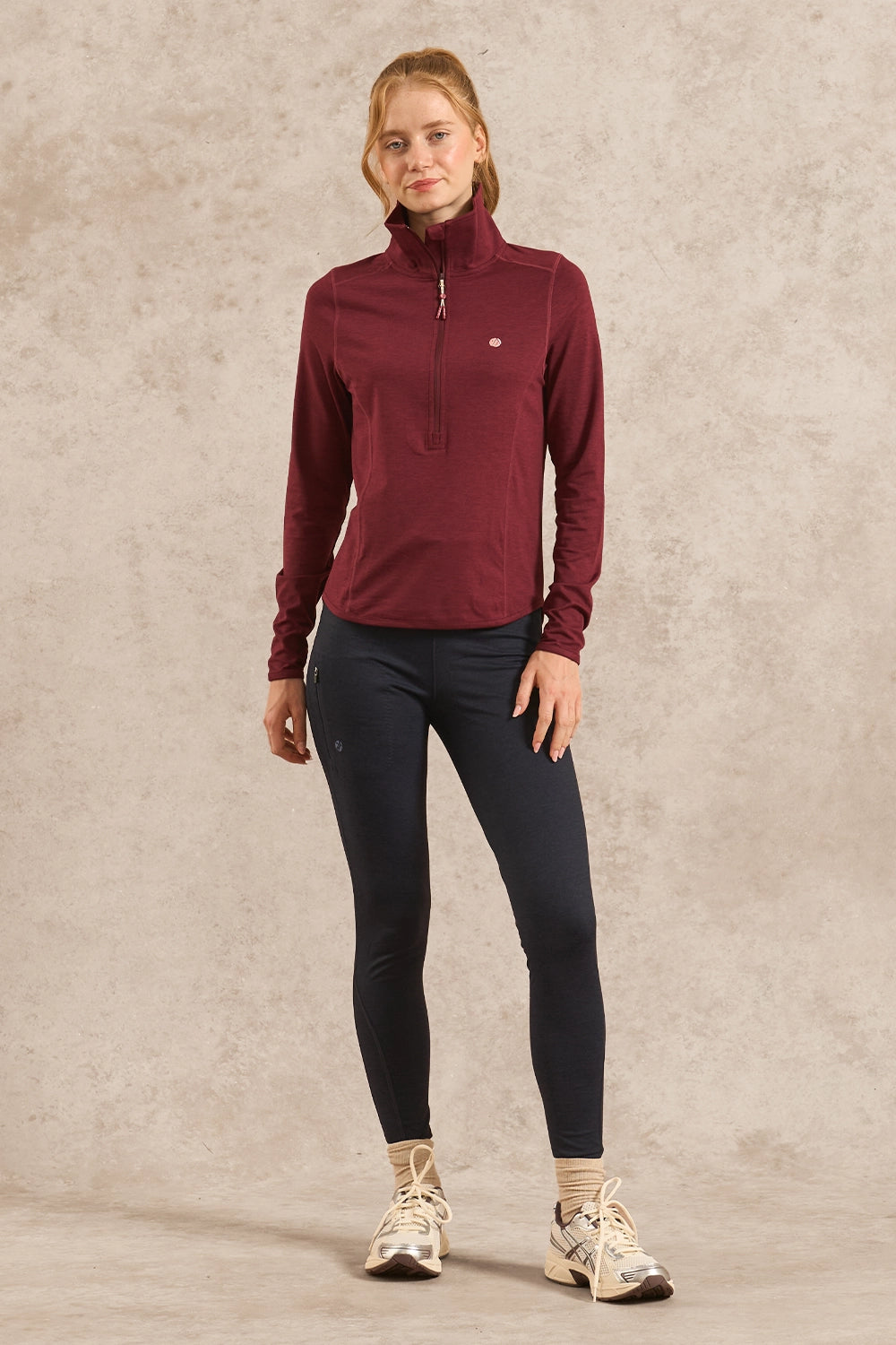 A woman modeling a maroon long-sleeve quarter-zip athletic pullover with dark navy leggings and beige running shoes, standing against a textured beige studio backdrop.