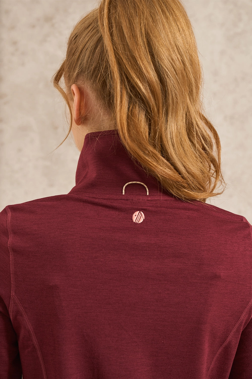 Back view of a woman with blonde hair in a ponytail wearing a heathered burgundy athletic quarter-zip jacket with a small circular logo on the back neck.