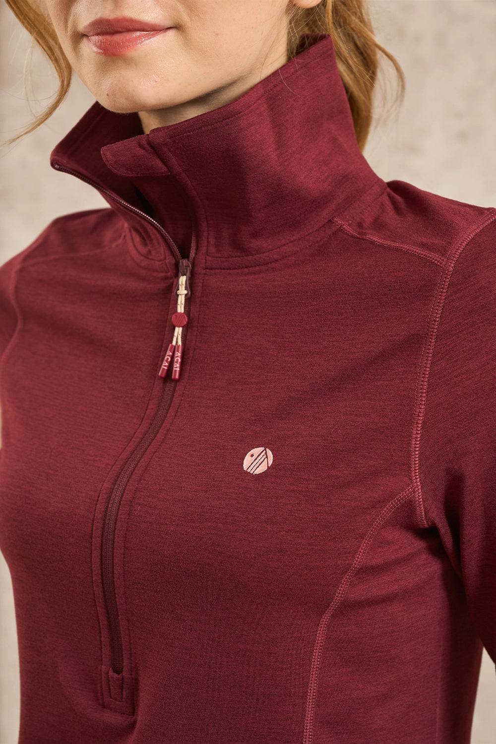 Close-up of a woman wearing a heathered burgundy half-zip athletic jacket with a high collar and a small circular logo on the chest.