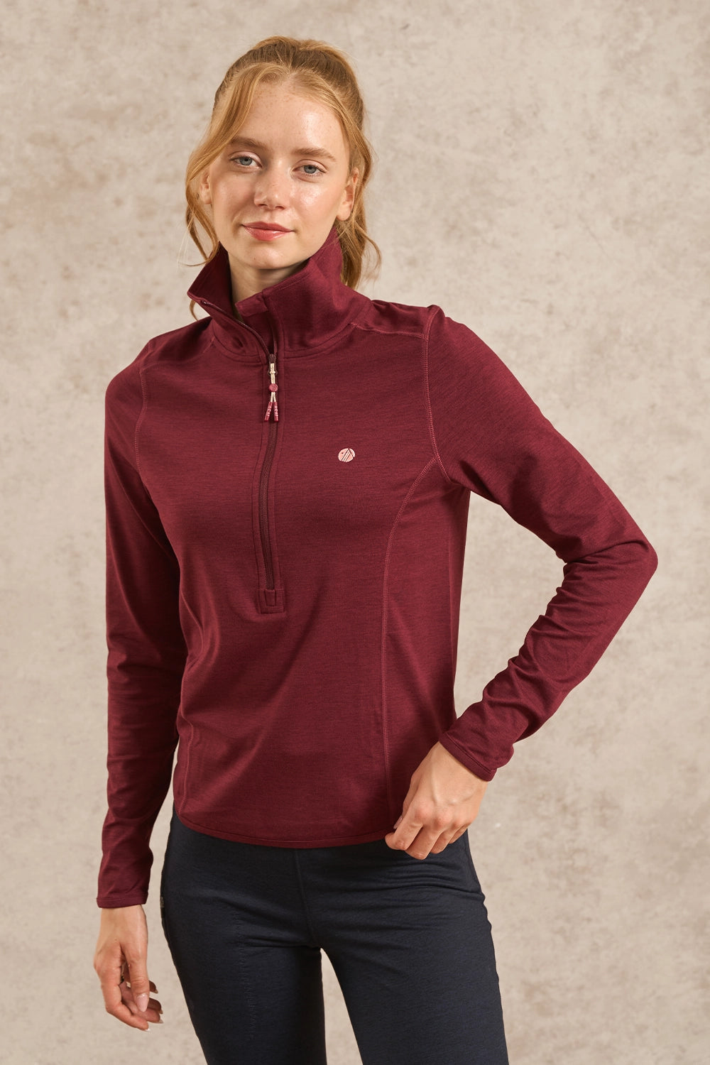 A woman wearing a burgundy long-sleeve quarter-zip pullover with a small circular logo on the left chest, paired with dark pants, standing against a textured beige background.