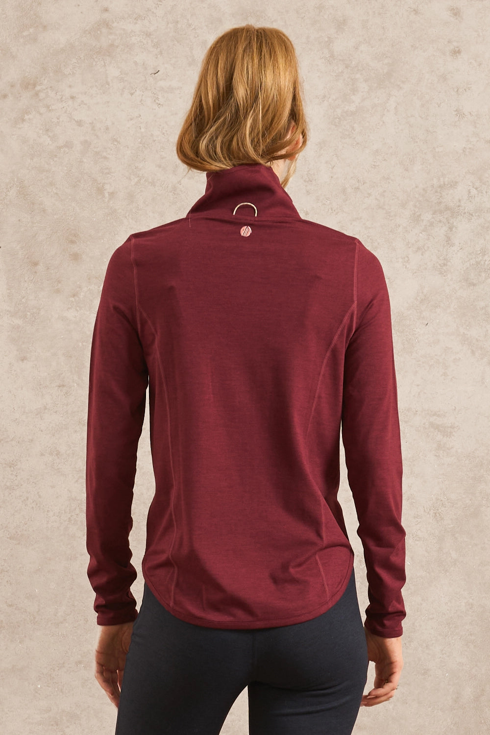 A woman viewed from behind wearing a burgundy long-sleeve mock neck athletic top with a small silver ring detail at the back of the collar, paired with dark leggings, against a textured beige background.