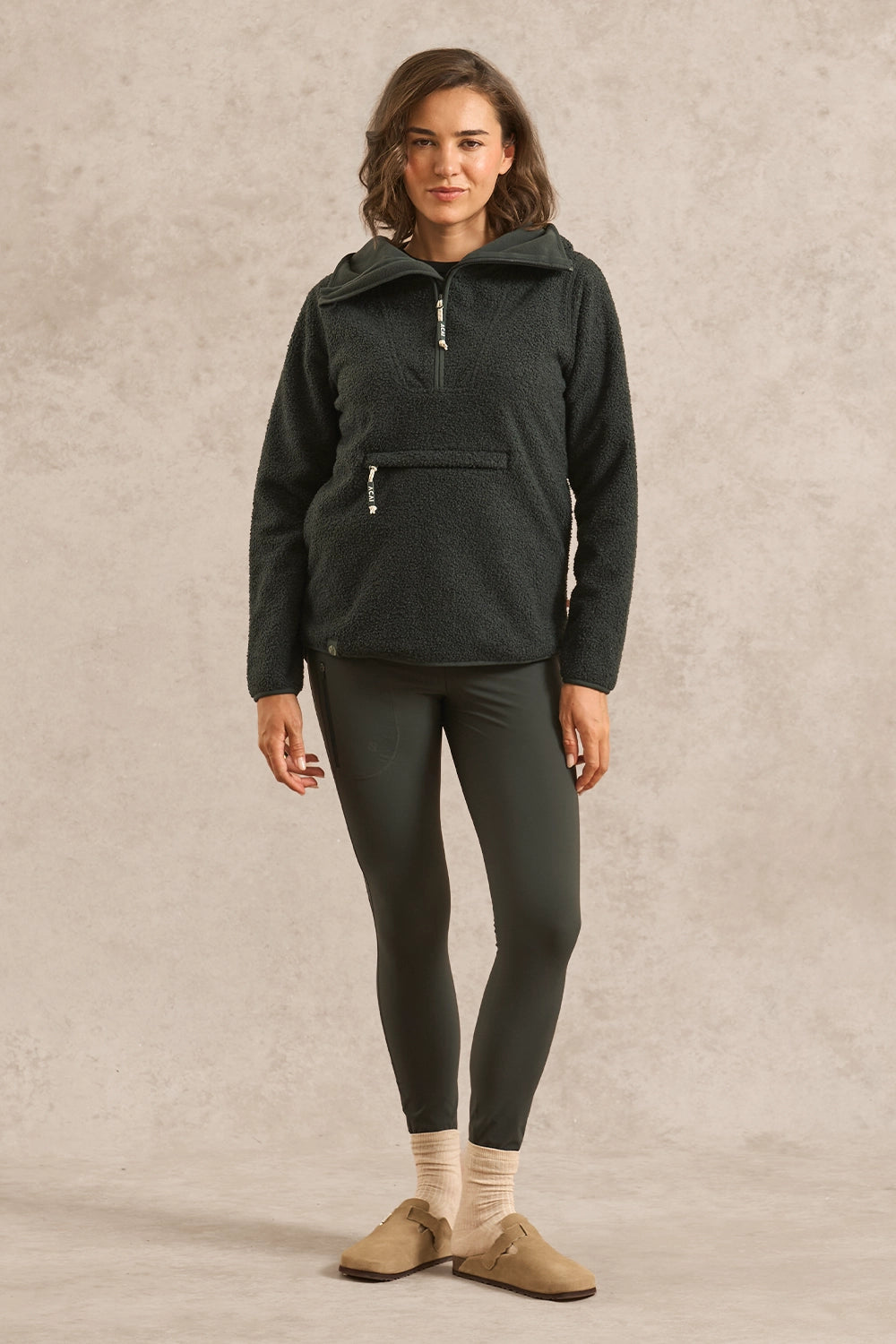 A woman stands against a textured beige studio backdrop wearing a dark forest green heathered pullover hoodie with a half-zip and zippered pocket, dark leggings, cream socks, and tan slip-on shoes.