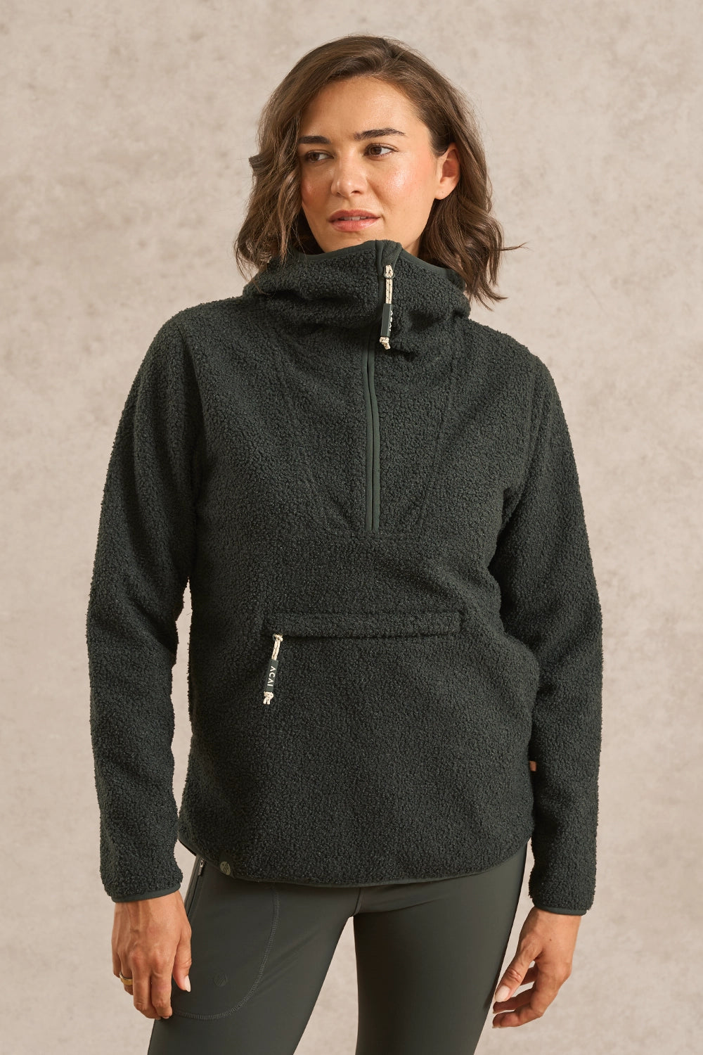 A woman wearing a forest green fleece pullover with a mock neck and a half-zip front, paired with dark gray leggings, standing against a textured beige wall.