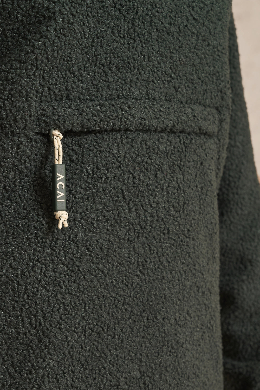 Close-up of a dark forest green fleece jacket featuring a front pocket with a drawstring closure. The drawstring has a black fabric tag with the white text 'ACAI' and a small white tassel.