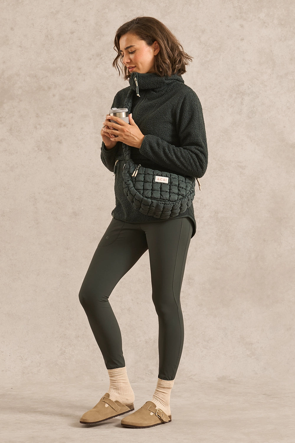 A woman stands against a textured beige studio background wearing a forest green fleece pullover, matching dark leggings, beige socks, and tan loafers. She is carrying a dark quilted crossbody bag and holding a metallic tumbler.
