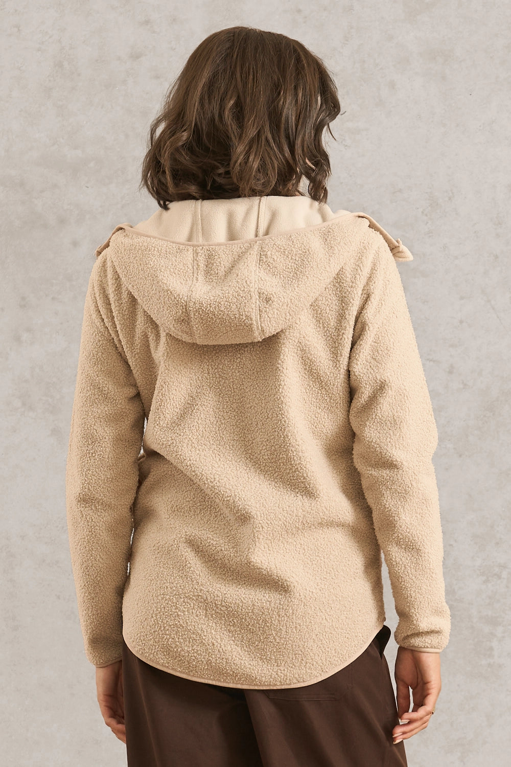 A person shown from the back wearing a beige sherpa fleece hooded jacket and dark brown pants, standing against a textured light gray wall.