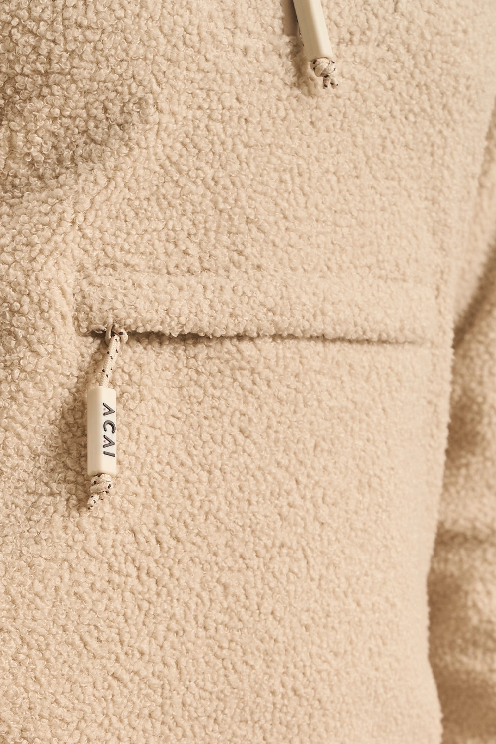 Close-up of a beige sherpa fleece jacket with a drawstring hood and a zippered pocket featuring a hanging cream pull tab printed with the brand name ACAI.