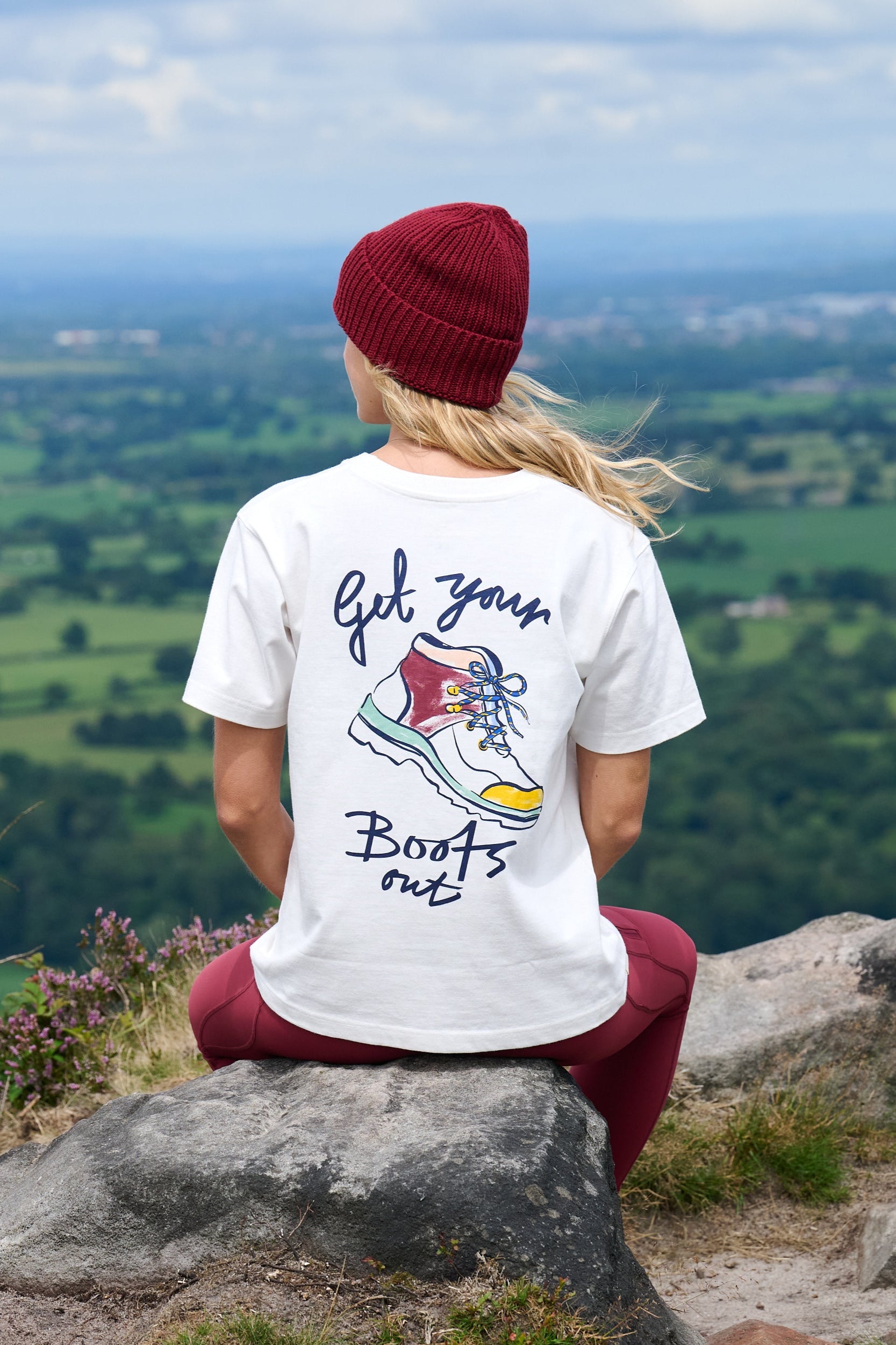 Person wearing a white t-shirt with 'Get your boots out' design, sitting on a rock overlooking a scenic landscape.