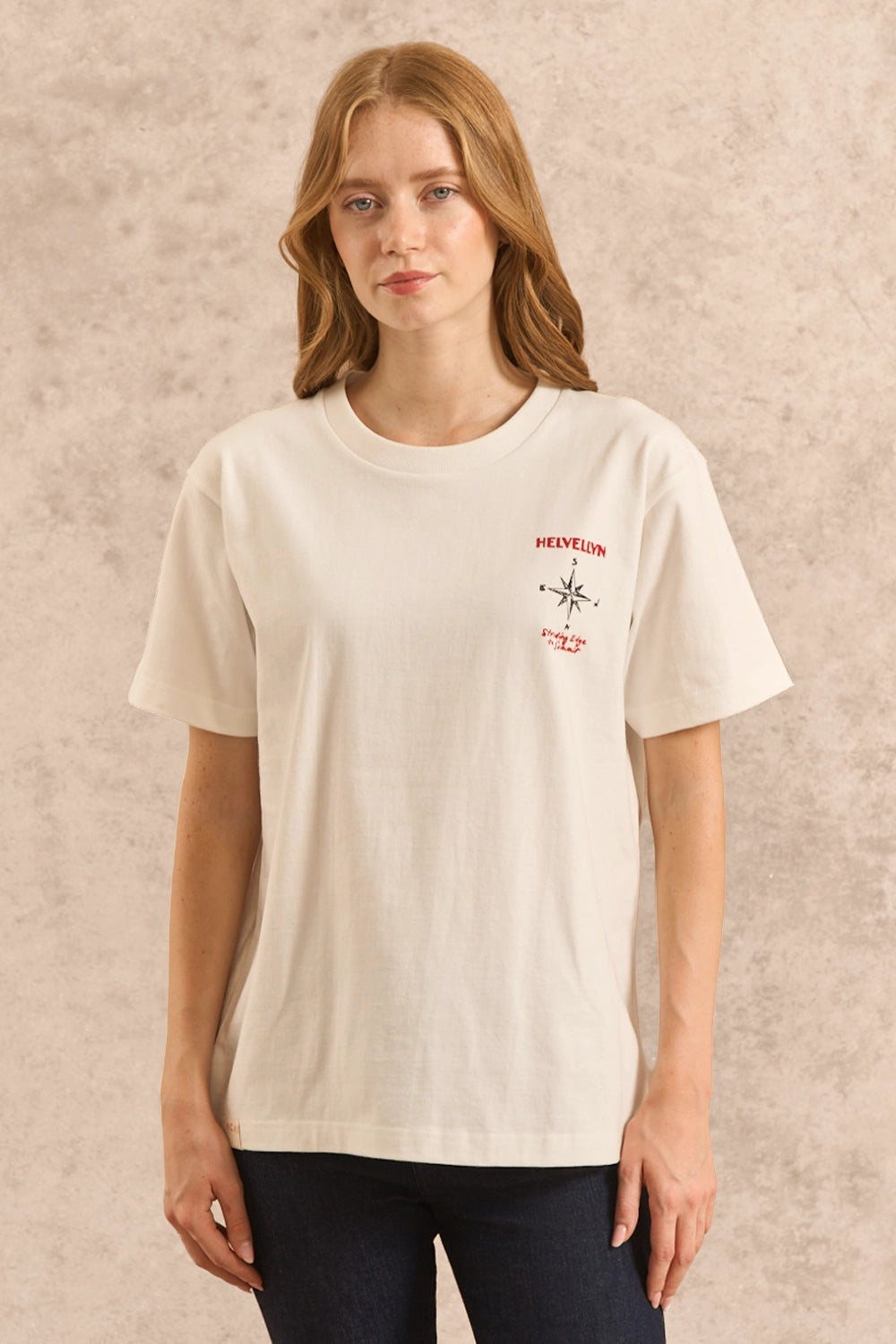 A woman modeling a cream-colored short-sleeve t-shirt with a small red and black compass graphic and 'HELVELLYN' text on the left chest, paired with dark blue jeans against a textured beige background.