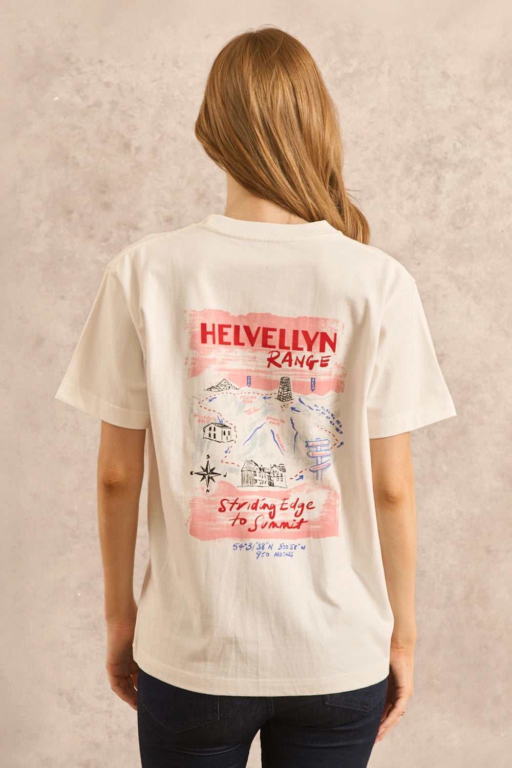 A woman viewed from behind wearing a cream-colored t-shirt with a pink and red graphic print on the back that reads 'HELVELYN RANGE' and 'Sf. Mary's Ridge', paired with dark blue jeans against a textured beige wall.
