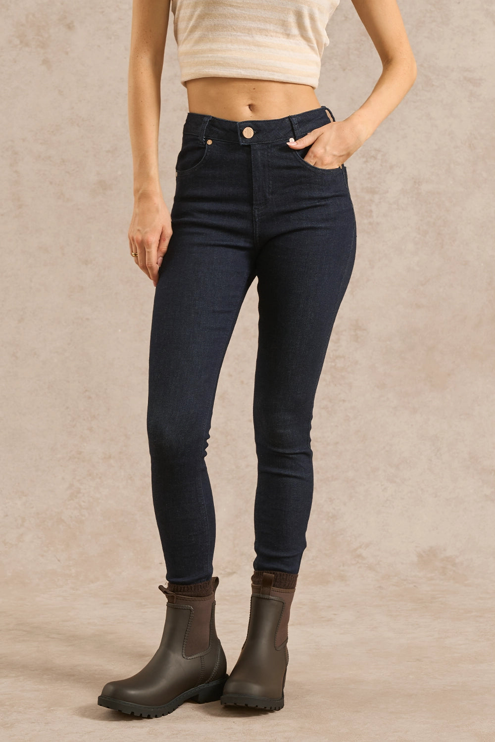 A model wearing dark wash skinny jeans and dark brown Chelsea boots, standing against a textured beige studio backdrop.
