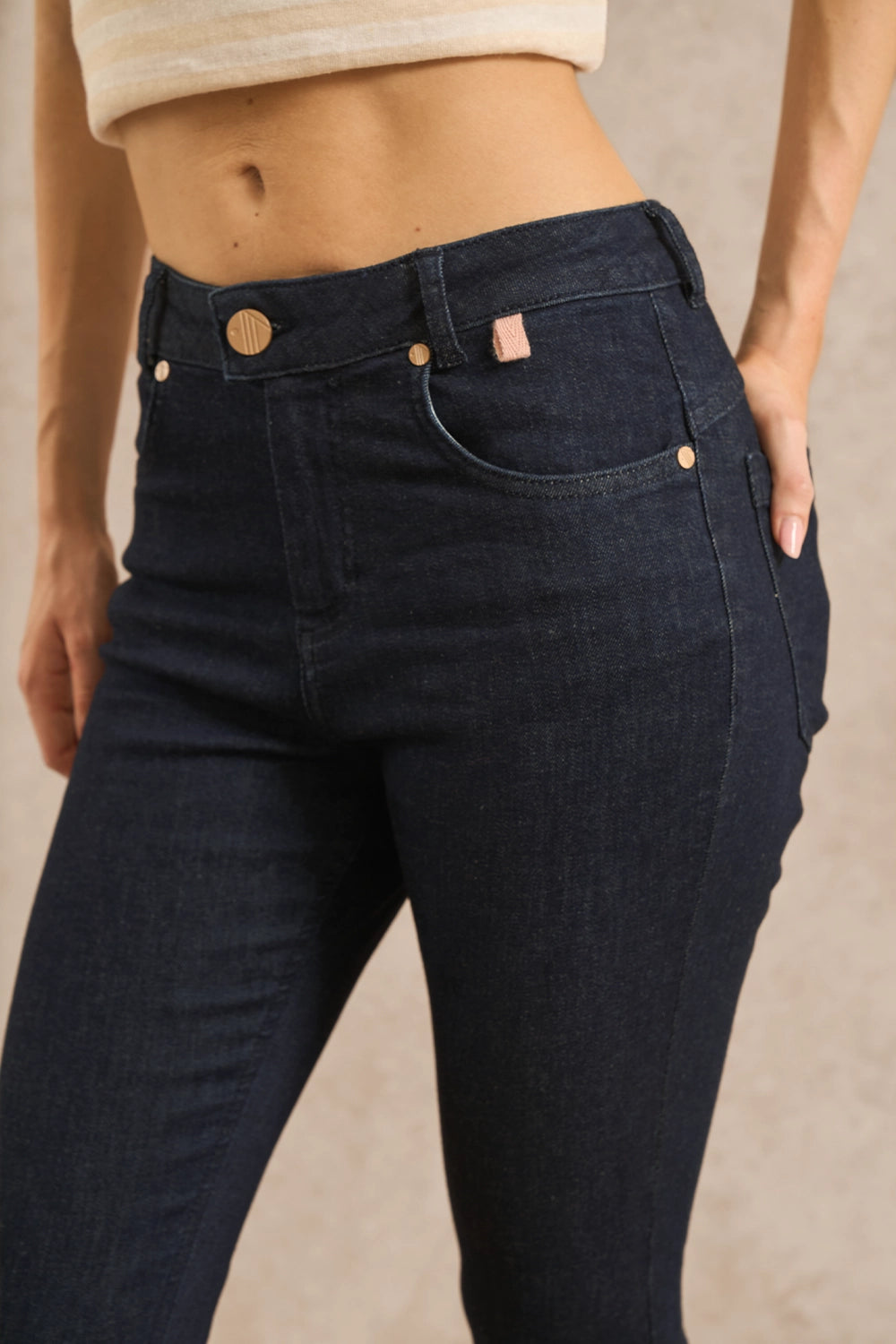 A close-up of a model wearing dark indigo skinny jeans with a small pink brand tab on the pocket, paired with a cream crop top against a neutral beige background.