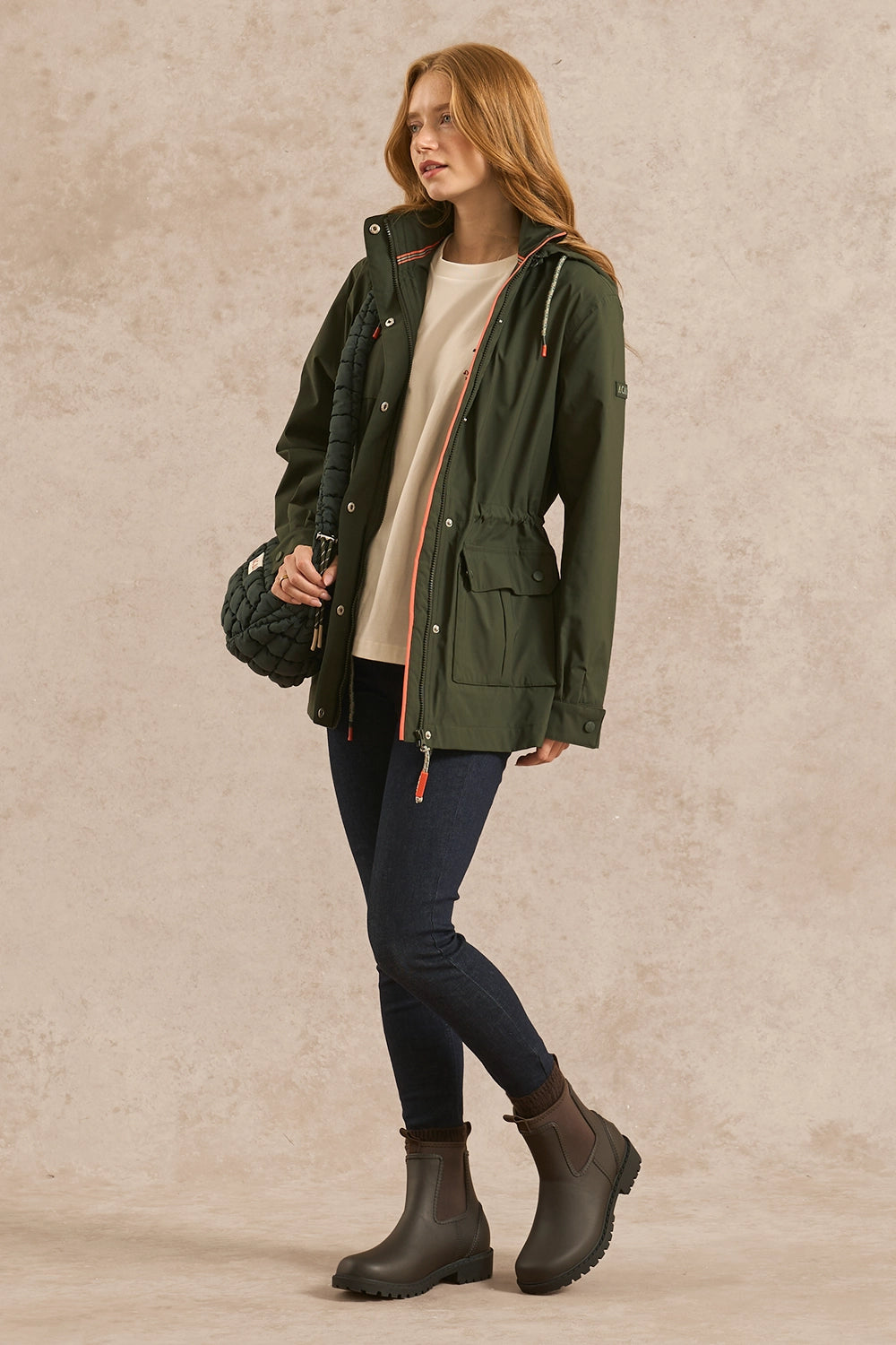 A woman models an olive green hooded parka jacket over a cream top, dark blue jeans, and dark brown boots, holding a black woven bag against a textured beige studio backdrop.