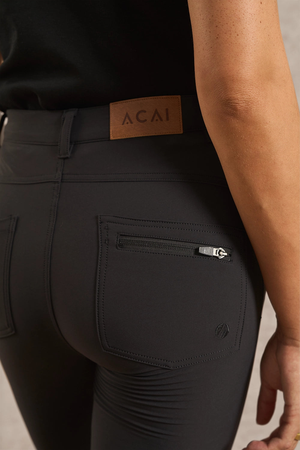 Close-up rear view of a person wearing black leggings with a brown leather Acai brand patch at the waistband and a zippered back pocket.