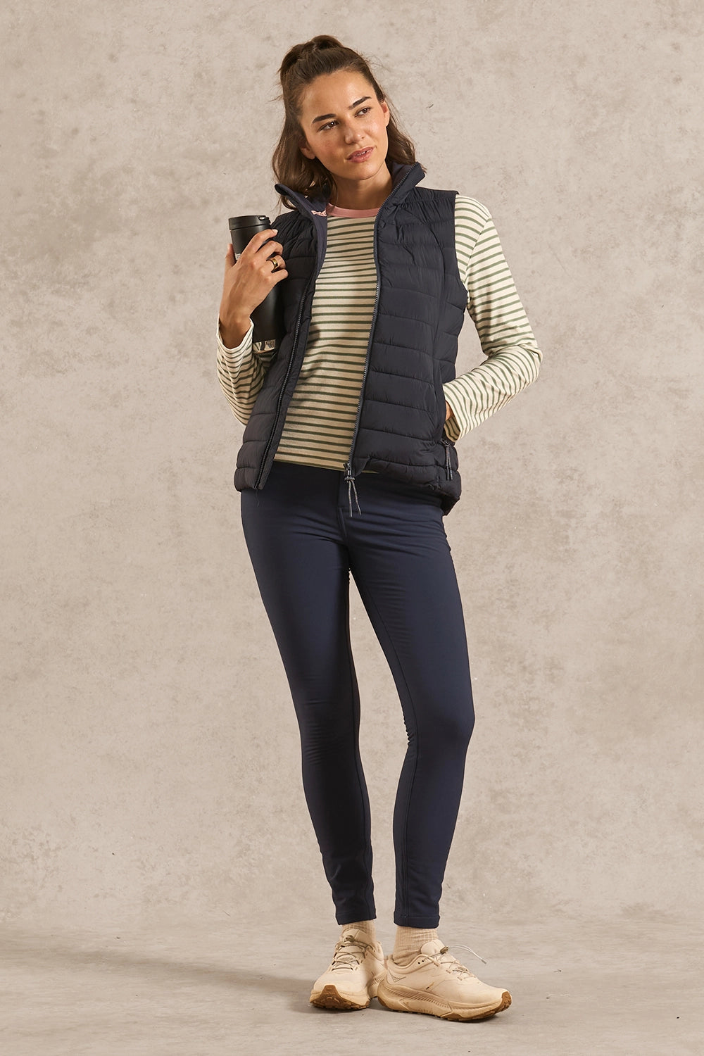 A woman modeling a navy blue sleeveless puffer vest over a cream and navy striped long-sleeve top, paired with navy leggings and beige sneakers, holding a black coffee mug against a textured beige studio backdrop.