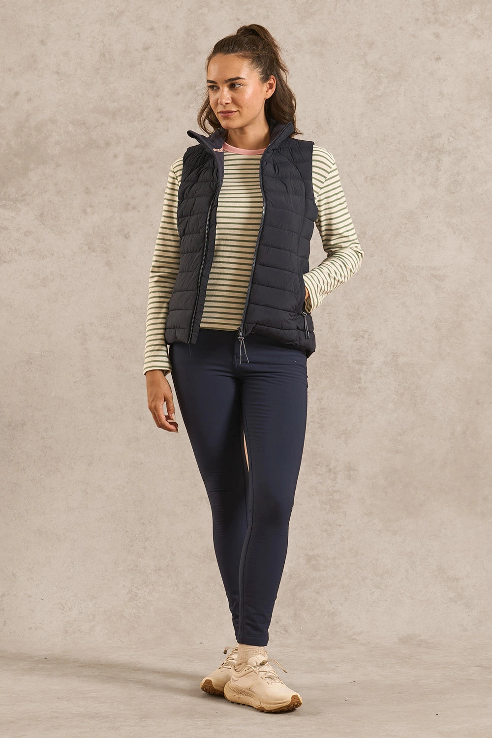 A woman modeling a sleeveless navy puffer vest over a cream and navy striped long-sleeve top, paired with navy leggings and beige sneakers, standing against a textured beige studio backdrop.