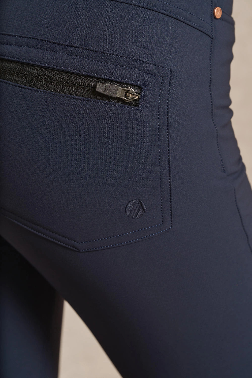 Close-up of navy blue leggings showing a zippered side pocket with a silver zipper pull and a small tonal logo near the hem.