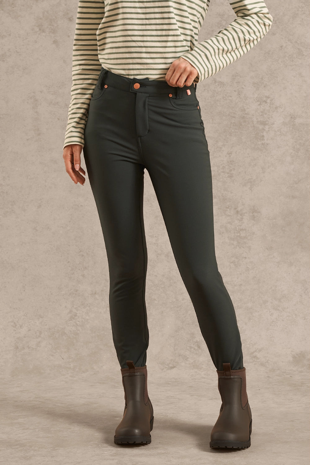 A model wearing dark olive green slim-fit riding breeches, a cream and green striped long-sleeve shirt, and dark brown Chelsea boots, standing against a textured gray studio backdrop.