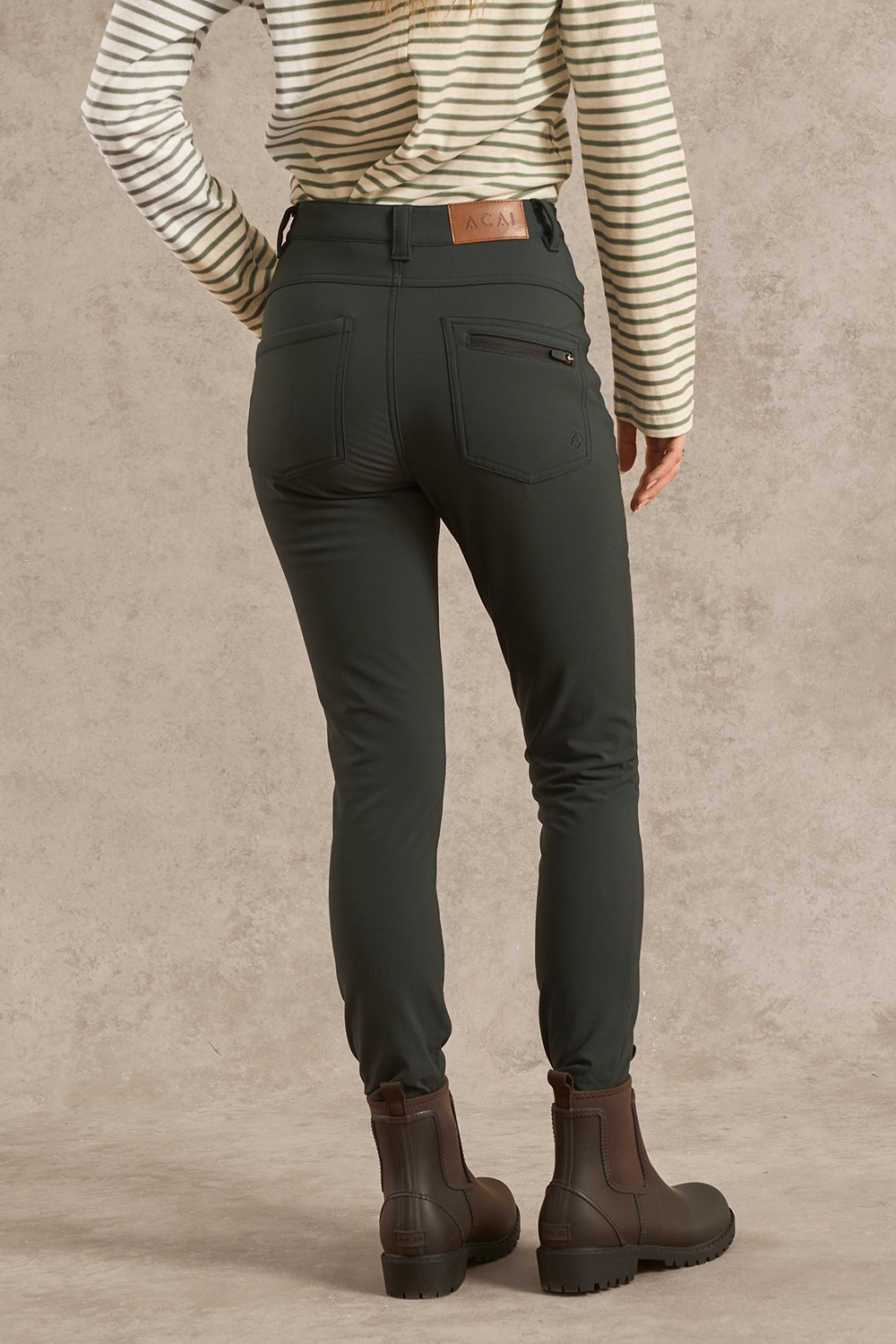 Rear view of a person wearing slim-fit dark olive green riding pants, a cream and brown striped long-sleeve shirt, and dark brown leather riding boots against a textured beige studio backdrop.