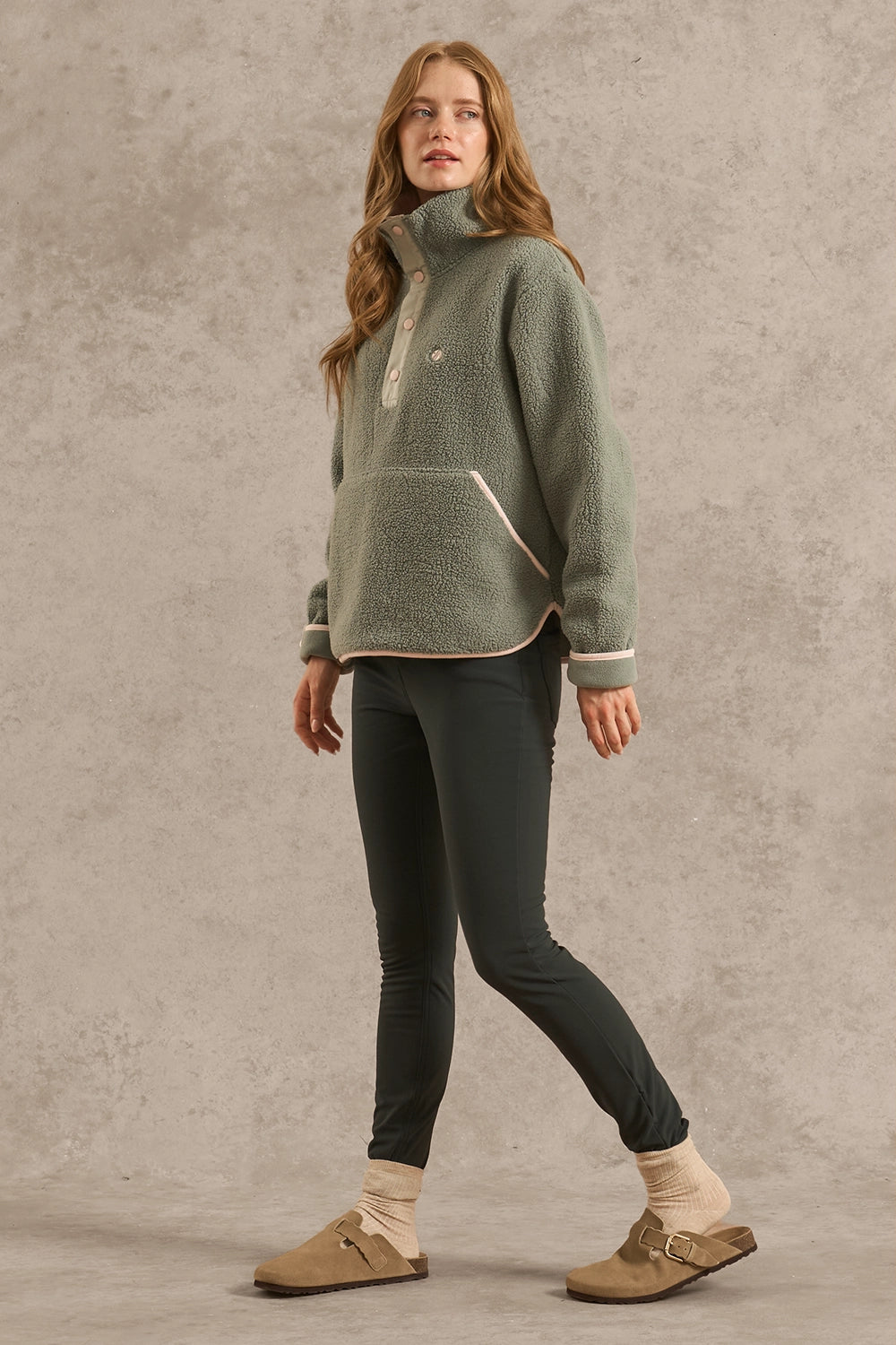 A woman modeling a sage green fleece pullover sweater with cream trim, paired with dark green leggings, beige socks, and brown mule sandals, set against a textured gray studio backdrop.